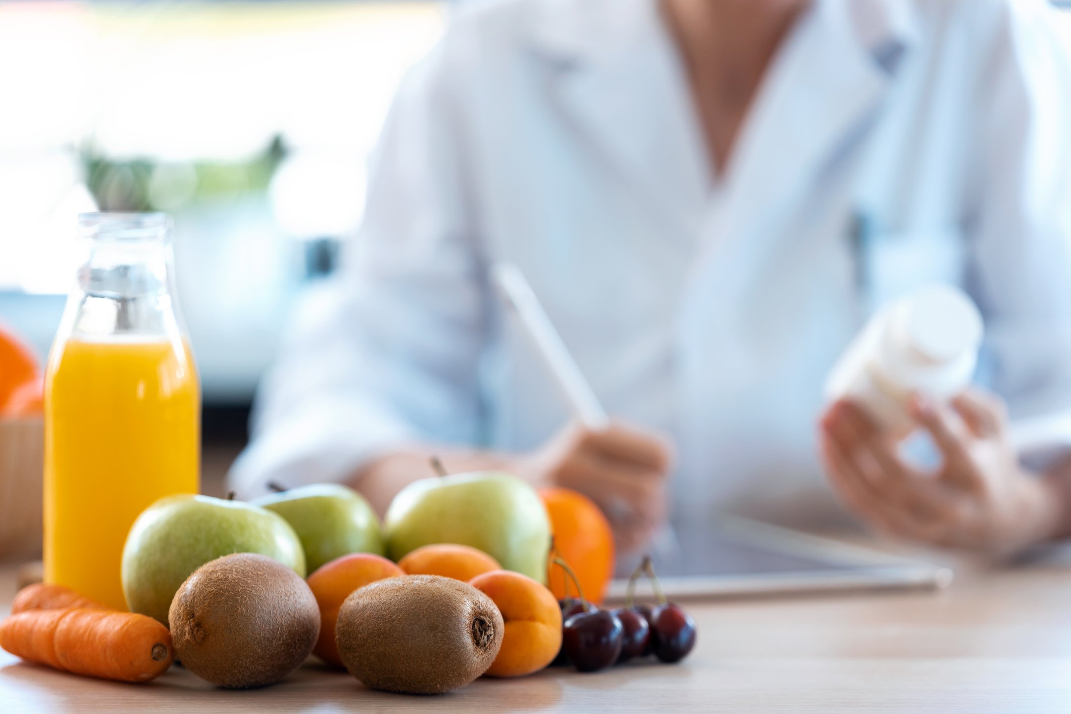 Fresh fruits, including apples, kiwis, carrots, apricots, cherries, and a bottle of orange juice on a kitchen counter, with a person in a white coat in the background.