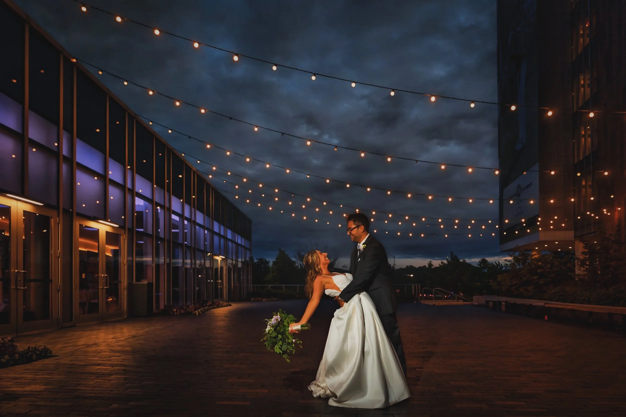 A bride and groom dancing outside at night under string lights, with modern glass building in the background.