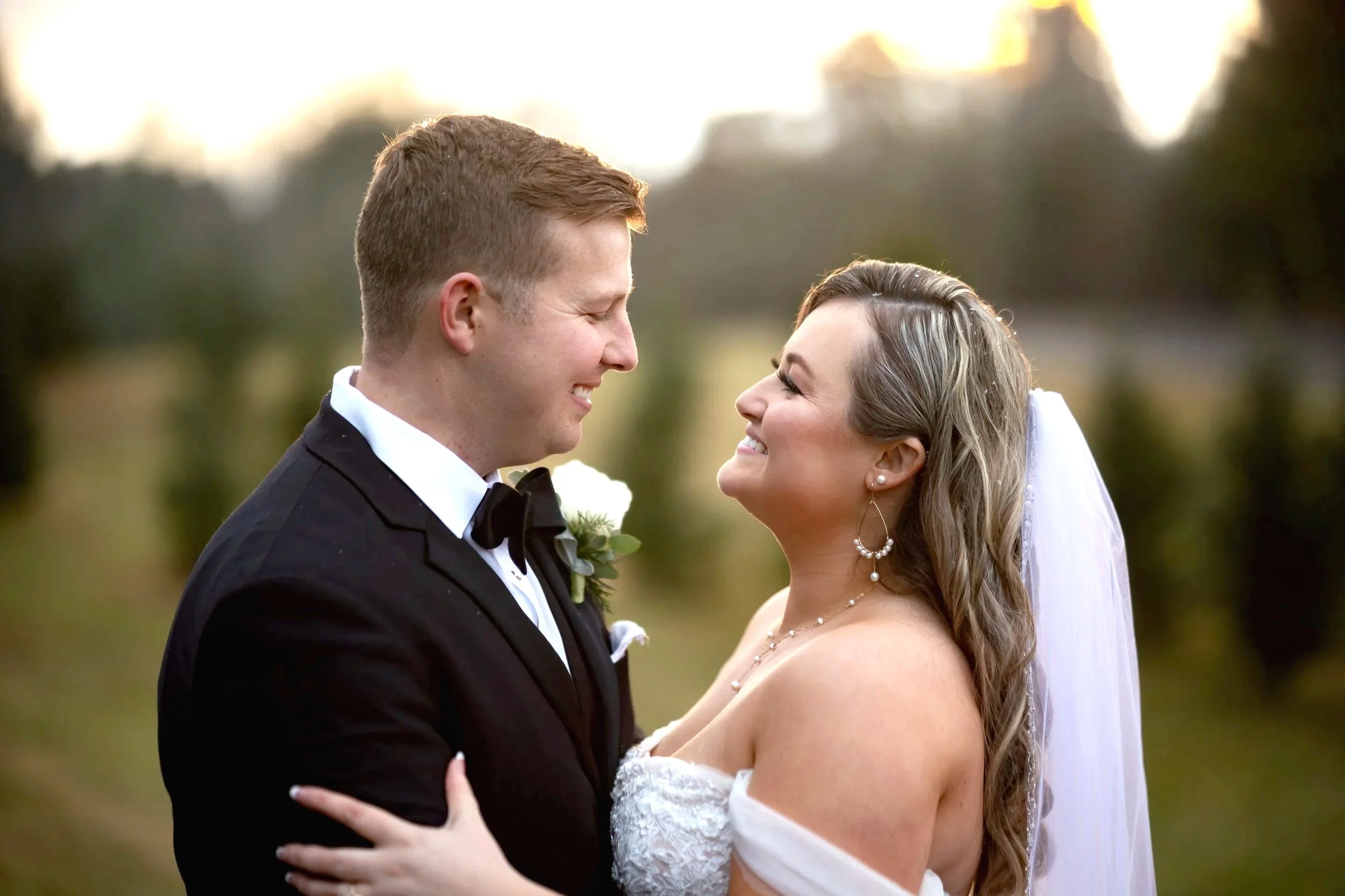 A newlywed couple smiling and looking into each other's eyes outdoors during sunset, with the groom in a black tuxedo and the bride in a white wedding dress and veil.