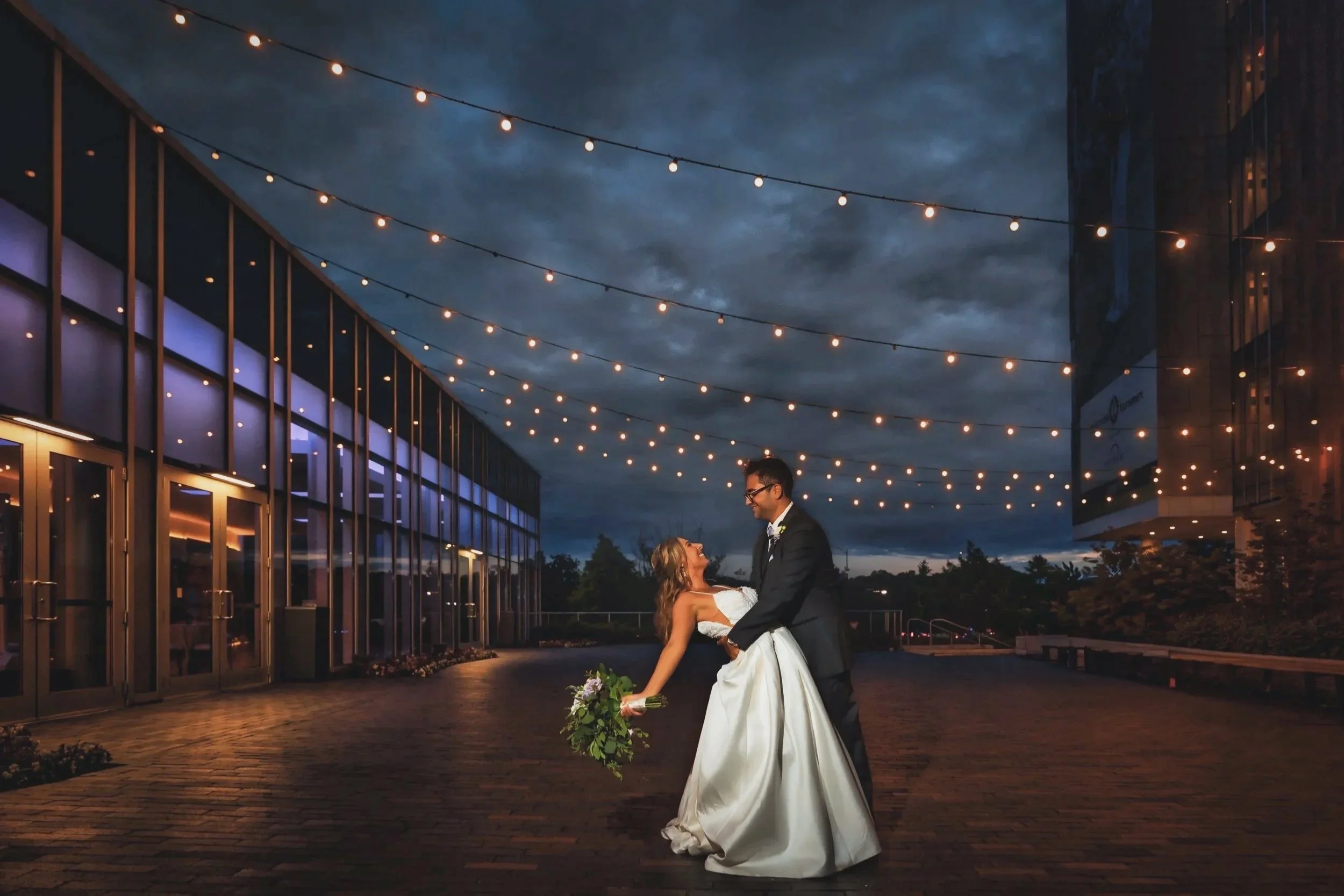 A bride and groom dancing outside at night under string lights, with modern glass building in the background.