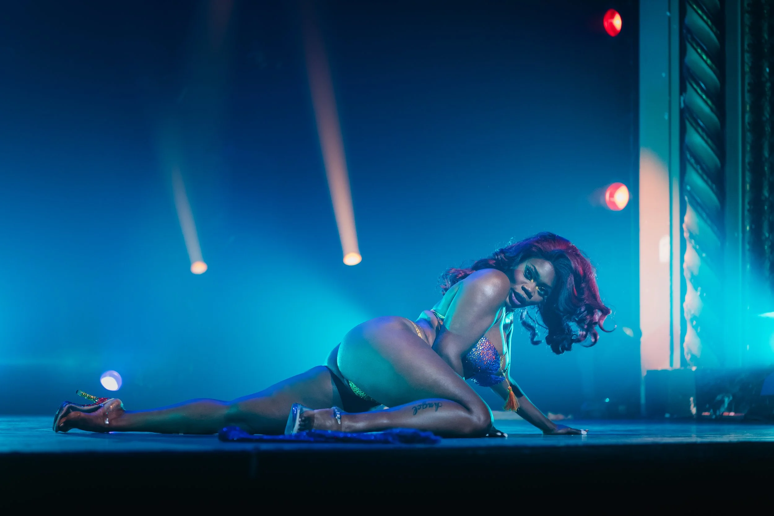 Foxy Lexi Brown with dark curly hair and purple lipstick poses on stage in a glittery outfit under blue and purple stage lighting.