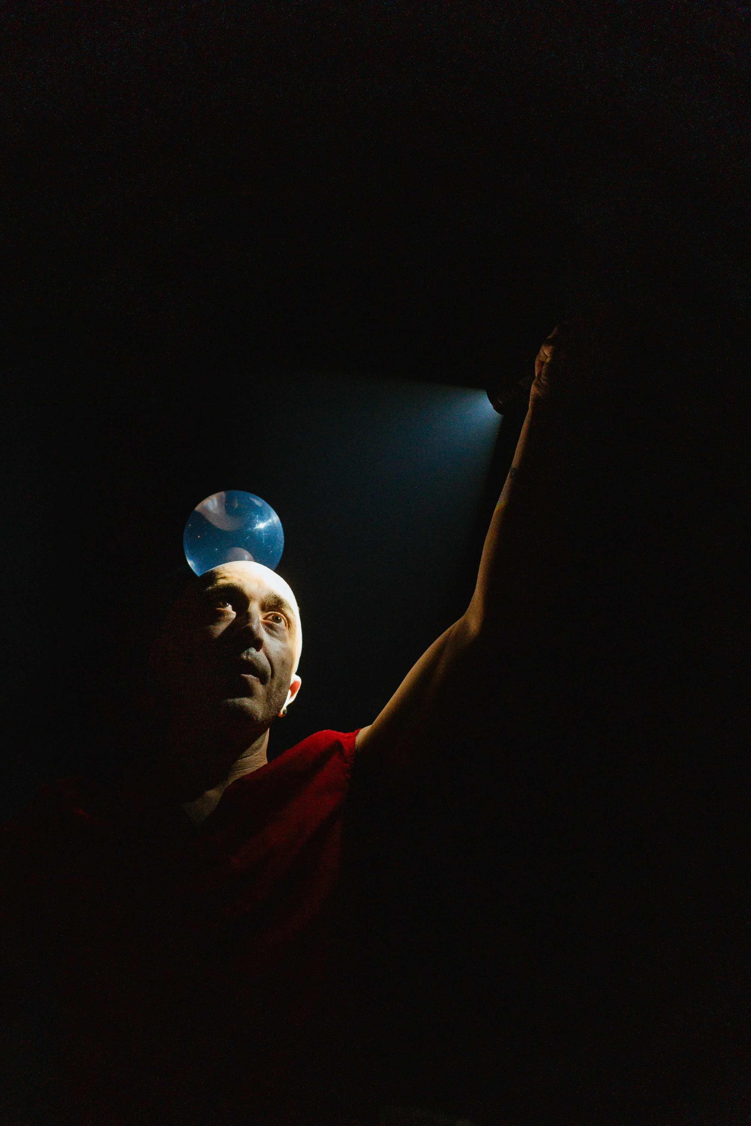 A performer on stage, balancing a glass ball on their head with a focused expression, illuminated by a spotlight against a dark background.