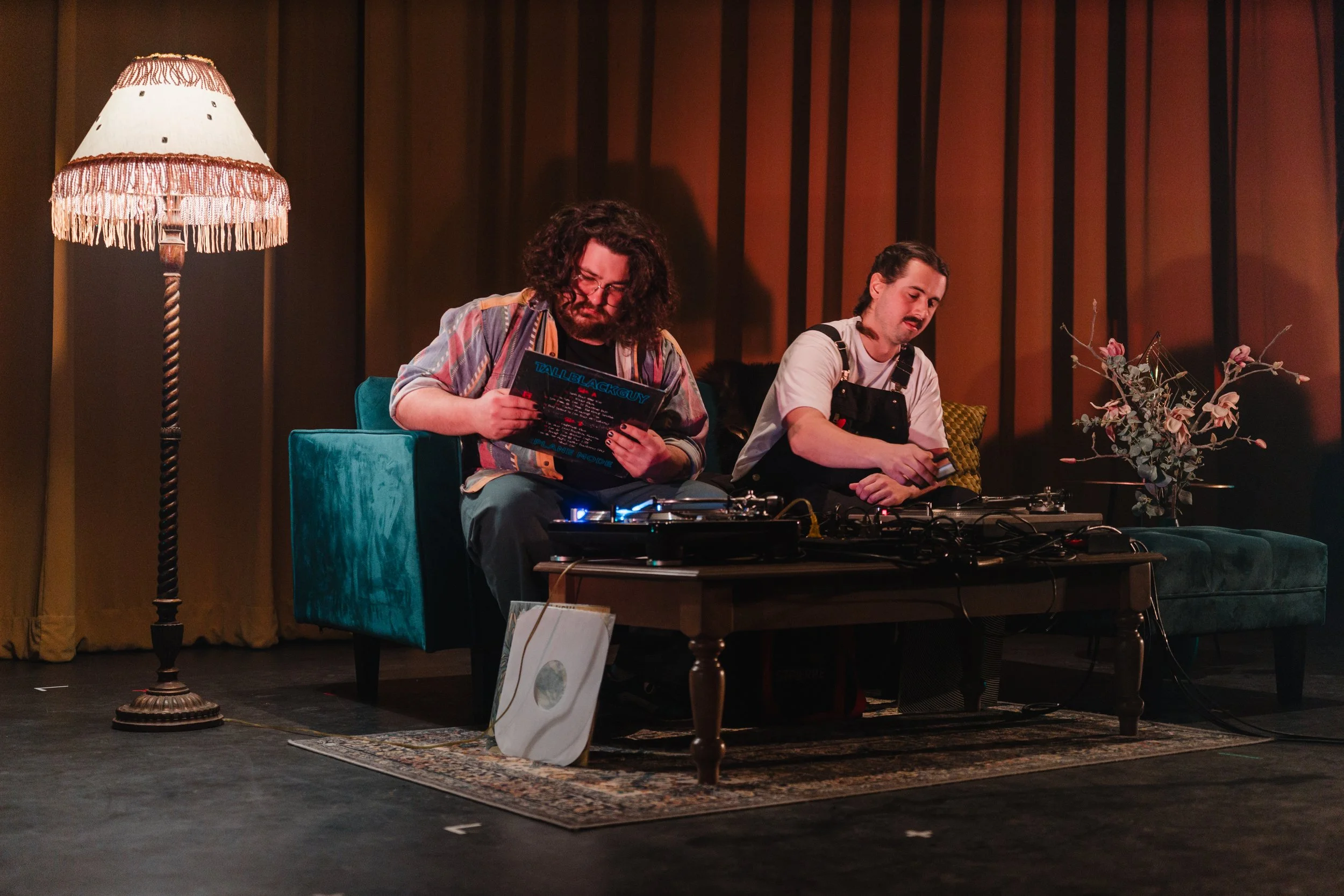 Two men sitting on a teal couch, one reading a vinyl record, the other using a turntable, in a cozy room with a tall lamp and a floral arrangement.