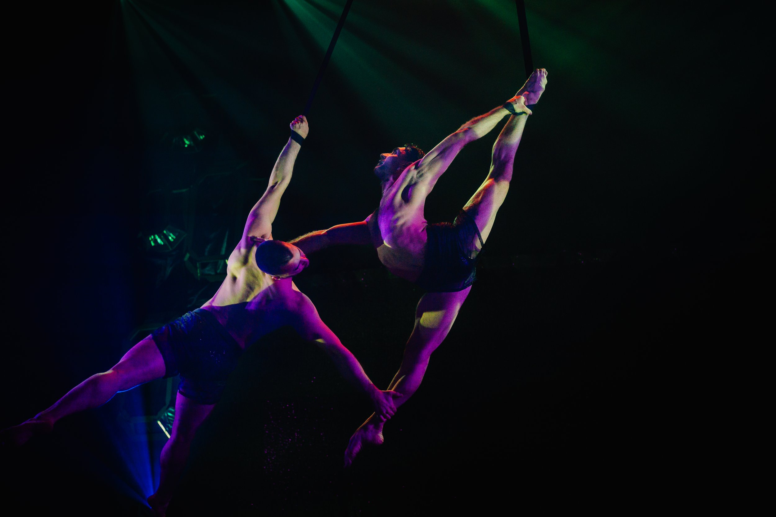Two male acrobats performing an aerial routine using a hoop, illuminated by colorful stage lights against a dark background.