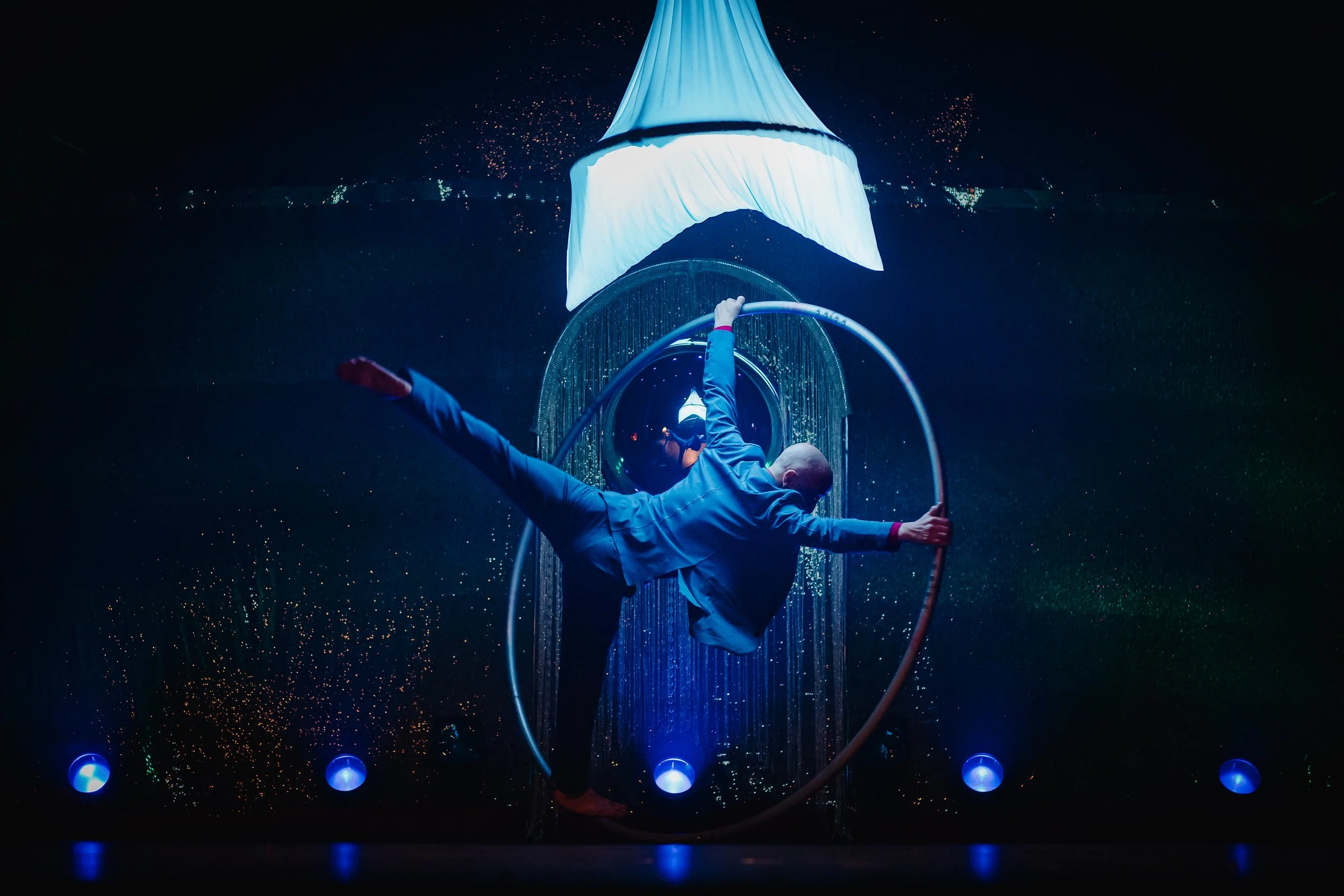 Performer in a blue outfit doing a contortion pose inside a metal hoop with a large illuminated fabric sculpture above, surrounded by dark background with colorful lights.