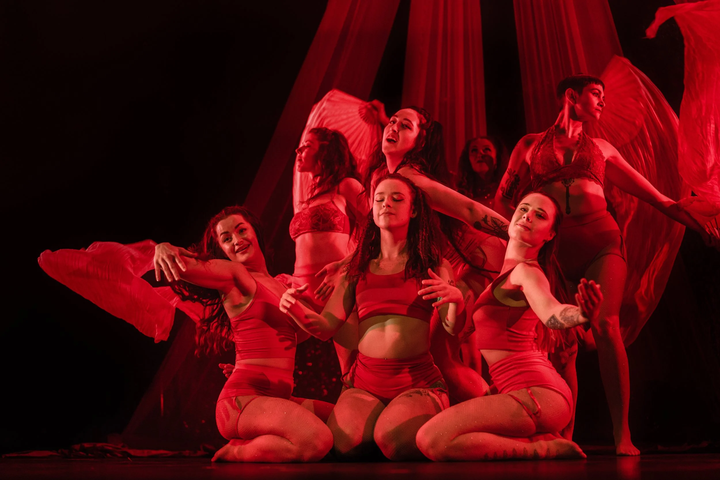 Group of seven female dancers performing on stage with red lighting and flowing fabric in the background.