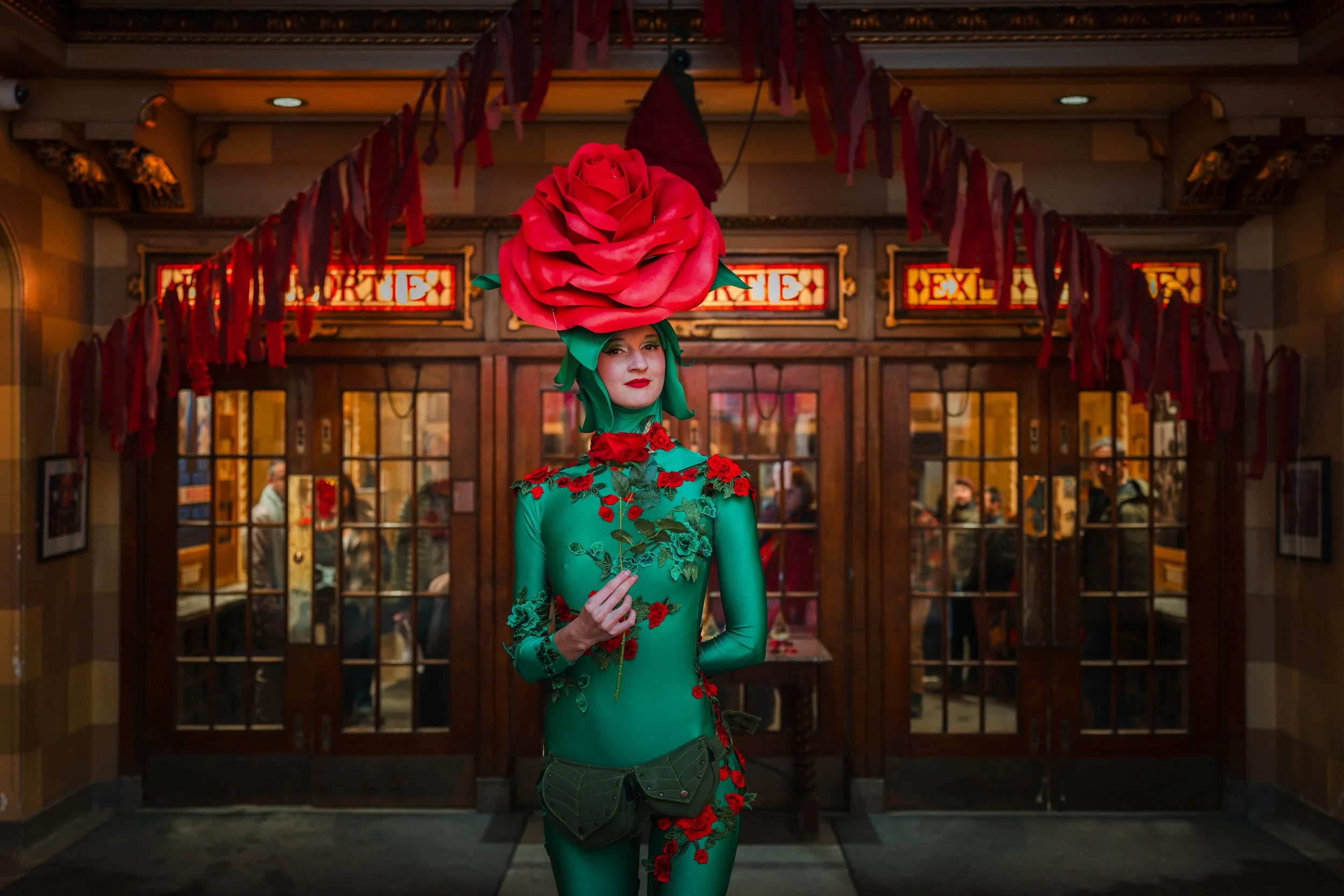 La rose de montreal in a green bodysuit with red floral embroidery and a large pink rose headpiece stands in front of the entrance to a restaurant, holding a small bouquet of red flowers.