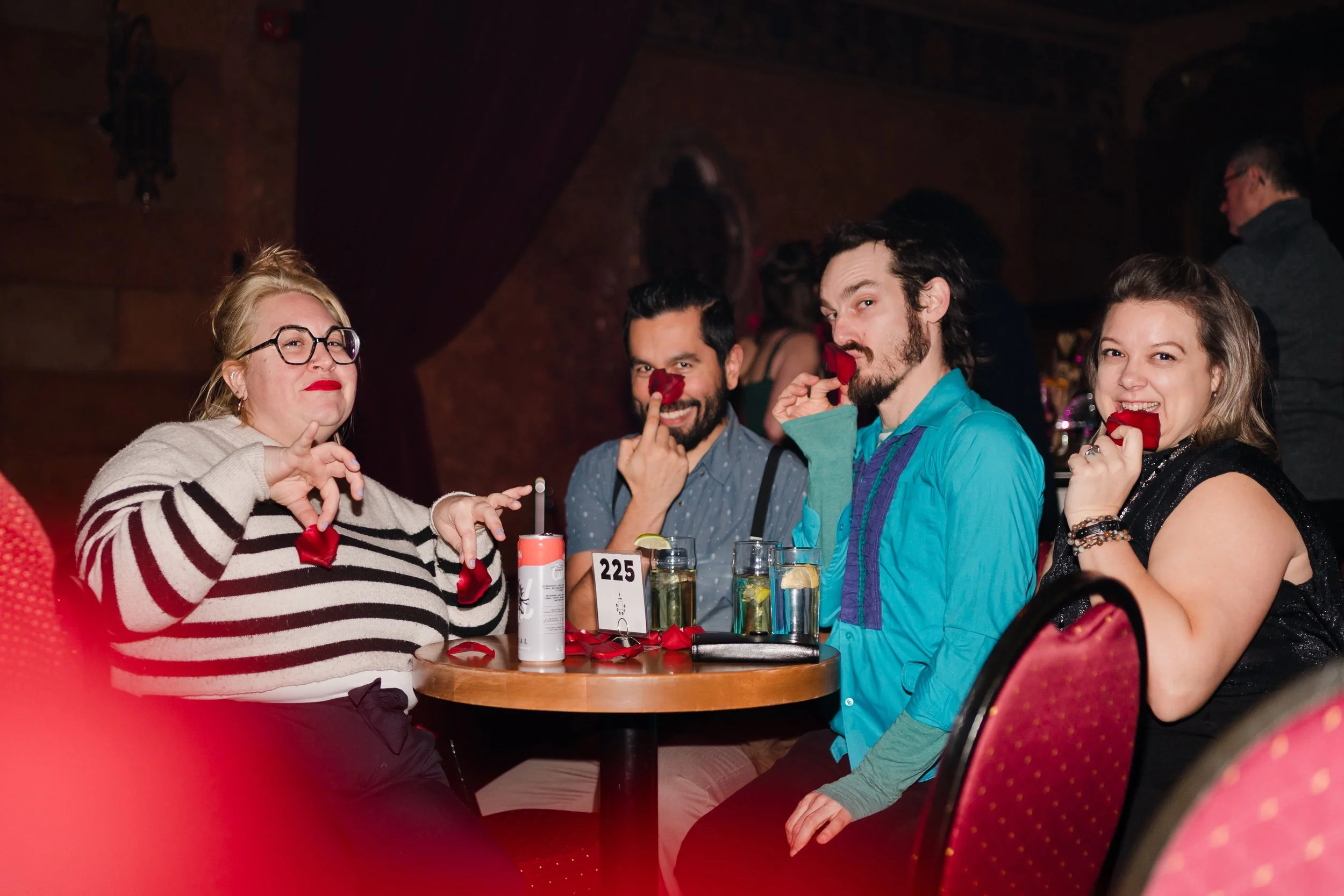 Four people sitting at a round table, each holding a red rose and making playful faces. The table has drinks, a number card '225', and rose petals scattered. The background shows a dimly lit, cozy indoor setting with other people and dark curtains.