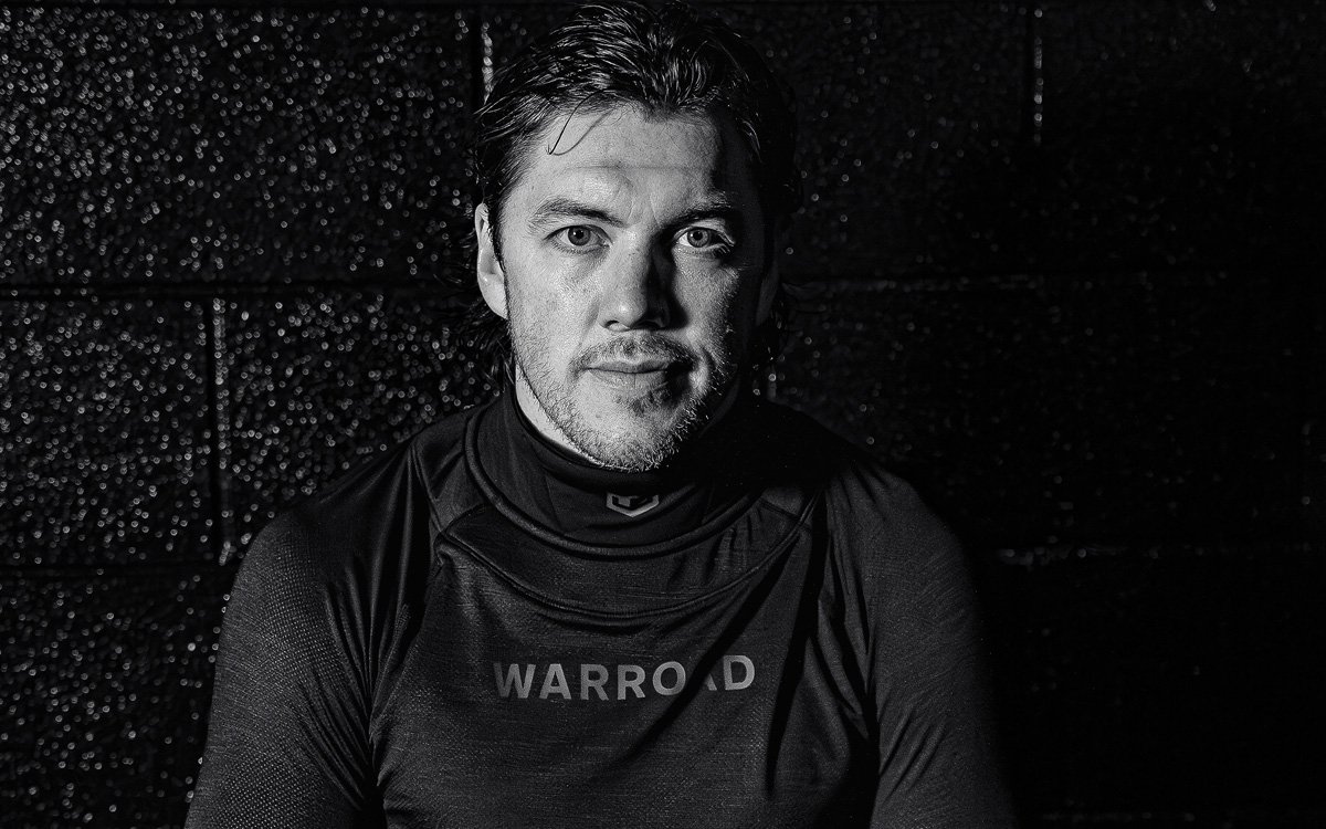 A black and white photo of TJ OSHIE wearing a TILO Neck Guard shirt with the word "WARROAD" on it, sitting against a dark, textured background. Warroad Hockey Co the most advanced cut-resistant technology in hockey - created by Sidfactor