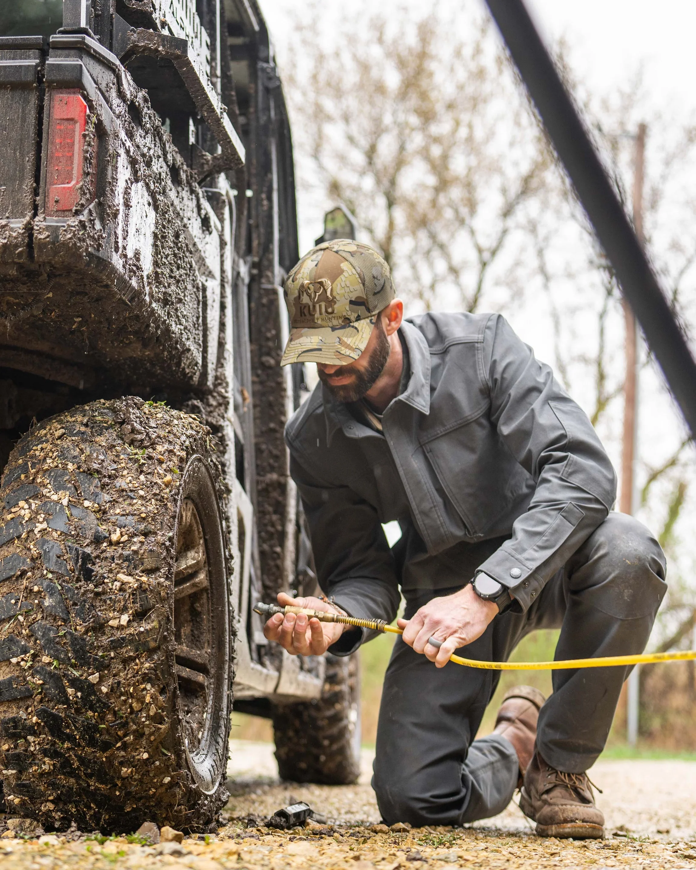 A man wearing a camouflage cap and gray jacket kneeling and working on the muddy tire of a large vehicle outdoors during daytime.  How do you translate the leading hunting companies DNA into lifestyle. Kuiu Lifestyle apparel created by Sidfactor