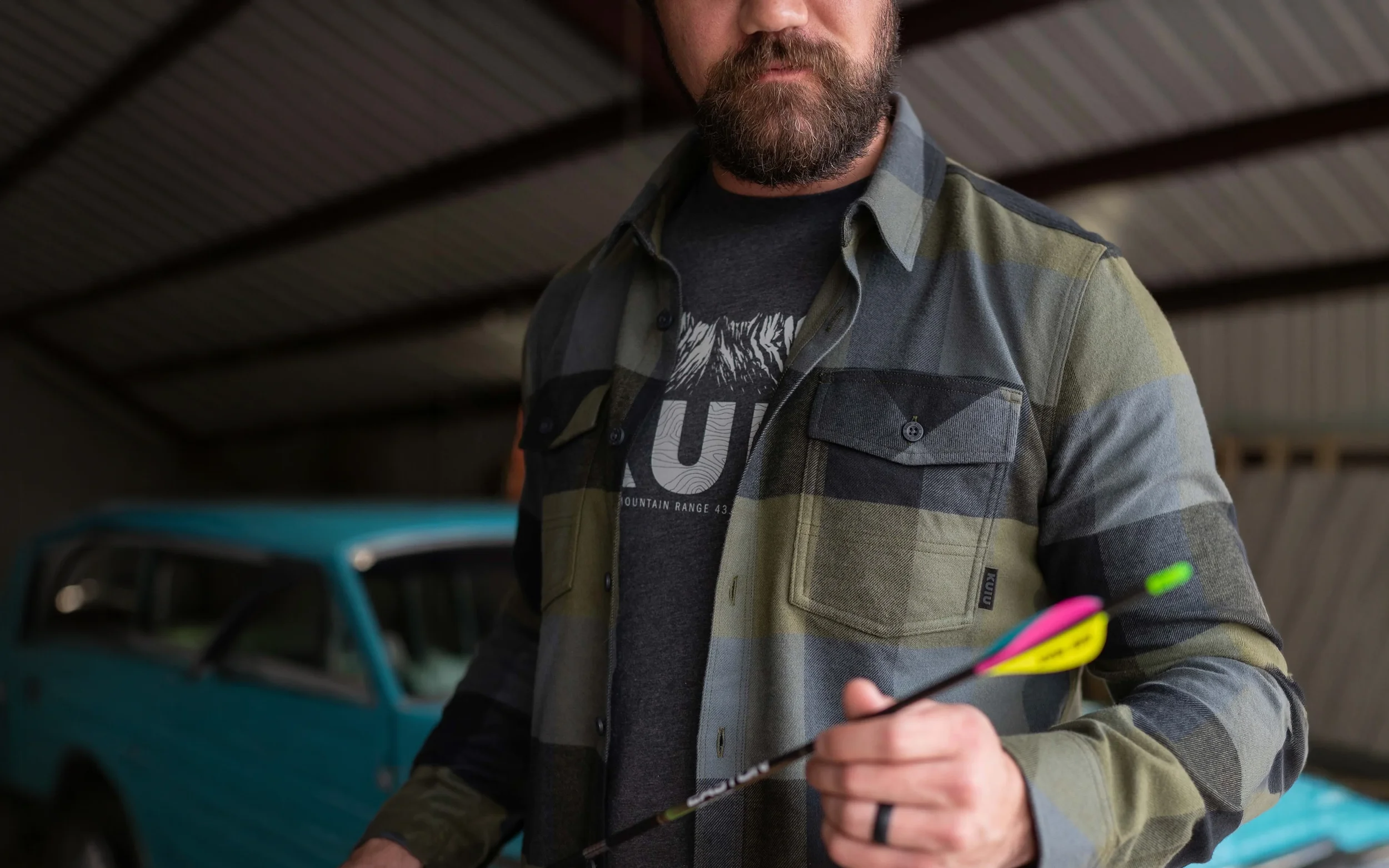 A man wearing KUIU with a beard wearing a green and gray plaid shirt over a black graphic T-shirt is holding arrows .   How do you translate the leading hunting companies DNA into lifestyle.  Kuiu Lifestyle apparel created by Sidfactor