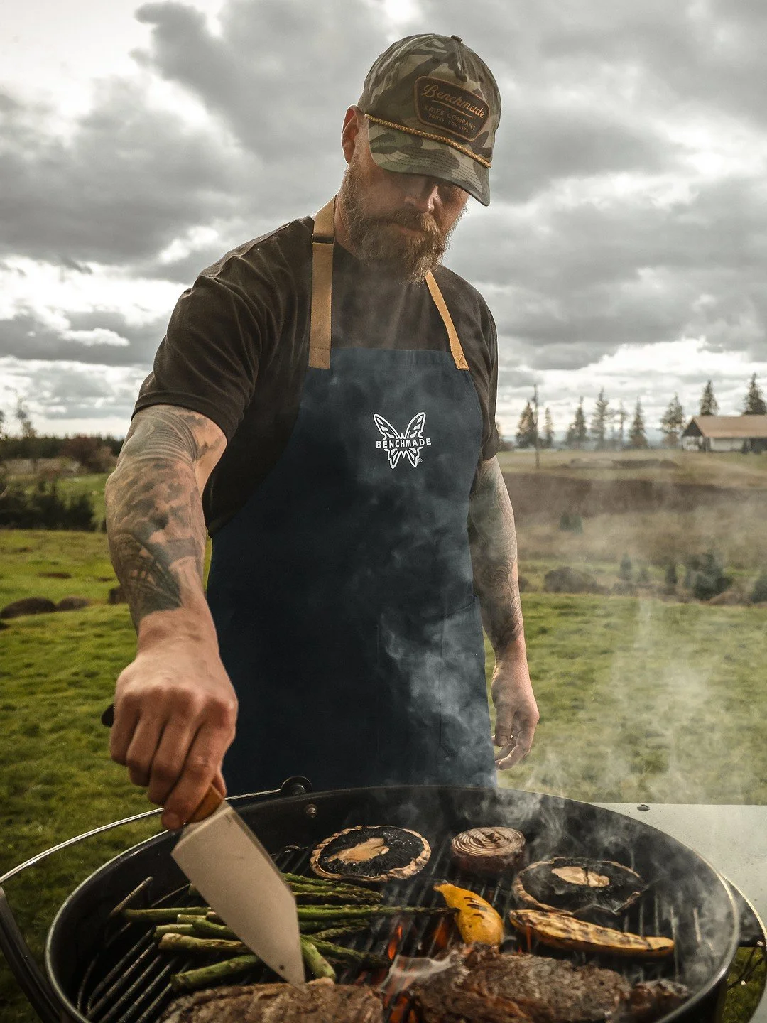 Man grilling outdoors, wearing a camouflage cap, black t-shirt, and apron with butterfly logo, cooking vegetables and meat on a charcoal barbecue under a cloudy sky.