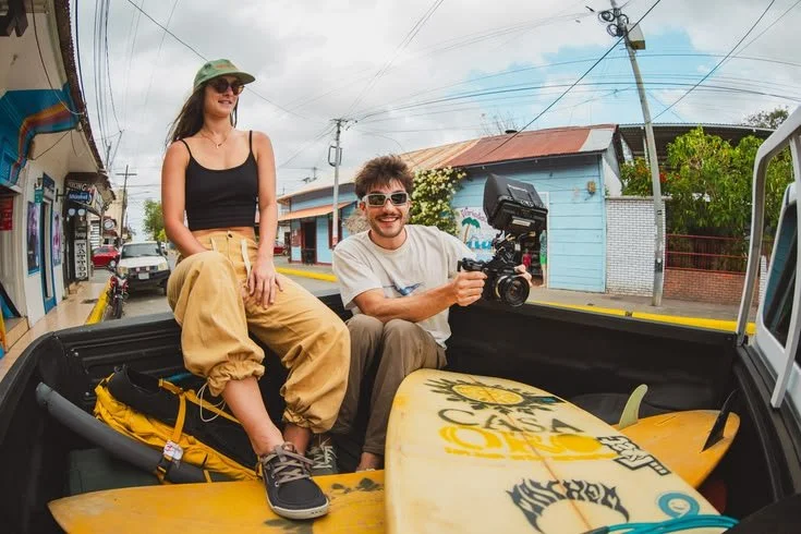 A woman and a man sitting in the back of a pickup truck with a surfboard, in a small town street, with buildings and power lines in the background.  Astral apparel created by Sidfactor