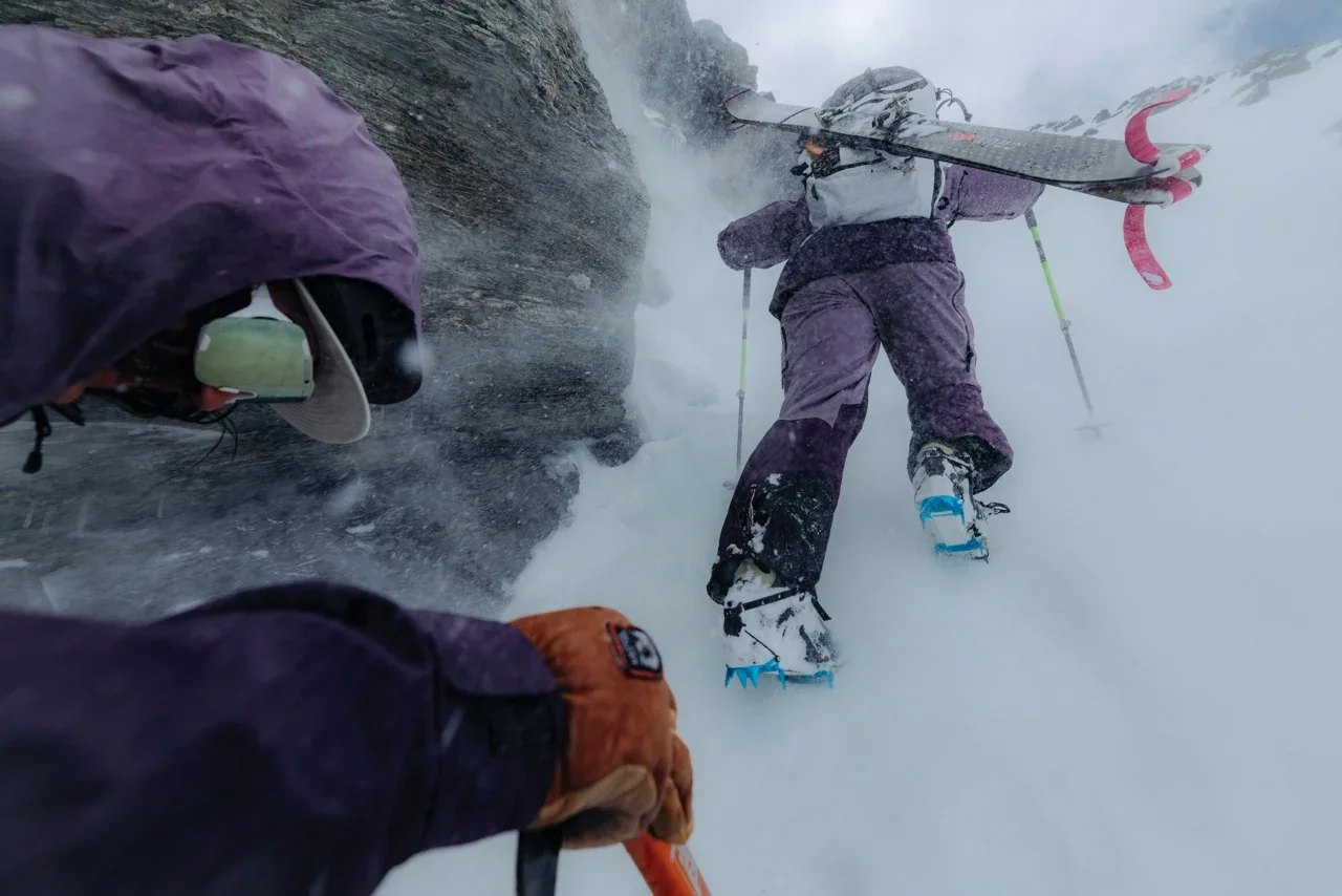 Two skiers ascending a snowy mountain slope with one carrying a snowboard on their shoulder and another wearing orange gloves, amidst falling snow and rugged terrain. Raide Research technical apparel created by Sidfactor