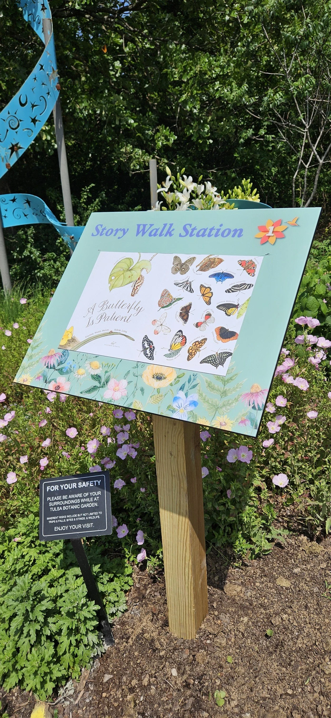 storywalk Sign at Tulsa Botanic Garden titled 'A Butterfly Is Patient,' featuring illustrations of various butterflies and a garden border with flowers, and a safety notice below informing visitors to be aware of their surroundings.