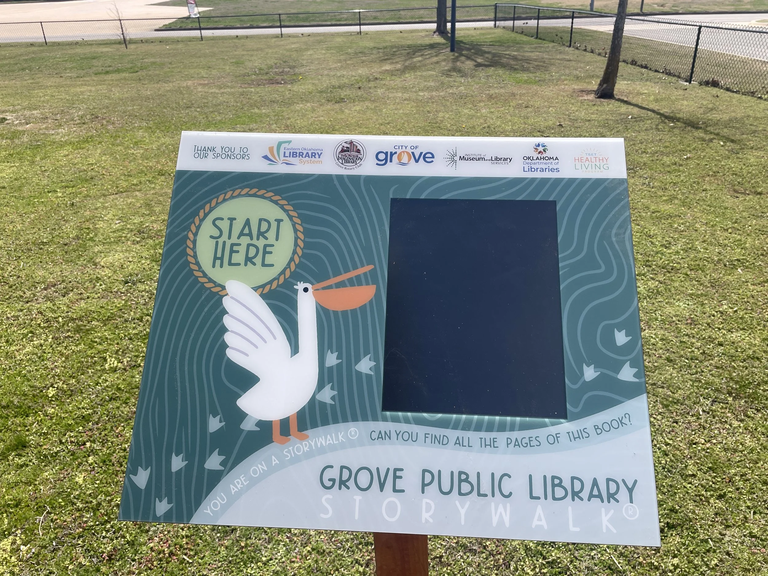 Story walk sign for Grove Public Library featuring a cartoon stork holding a book and the text "Start Here". The sign is outdoors on a grassy area with trees and a fence in the background.