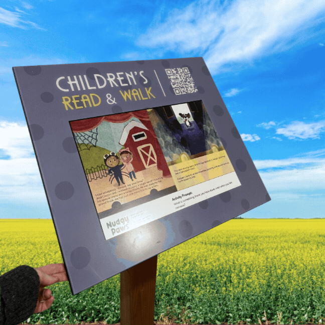 Children's story trail sign that opens. a signboard with colorful illustrations of a barn, animals, and children, mounted on a wooden post in a field under a blue sky.