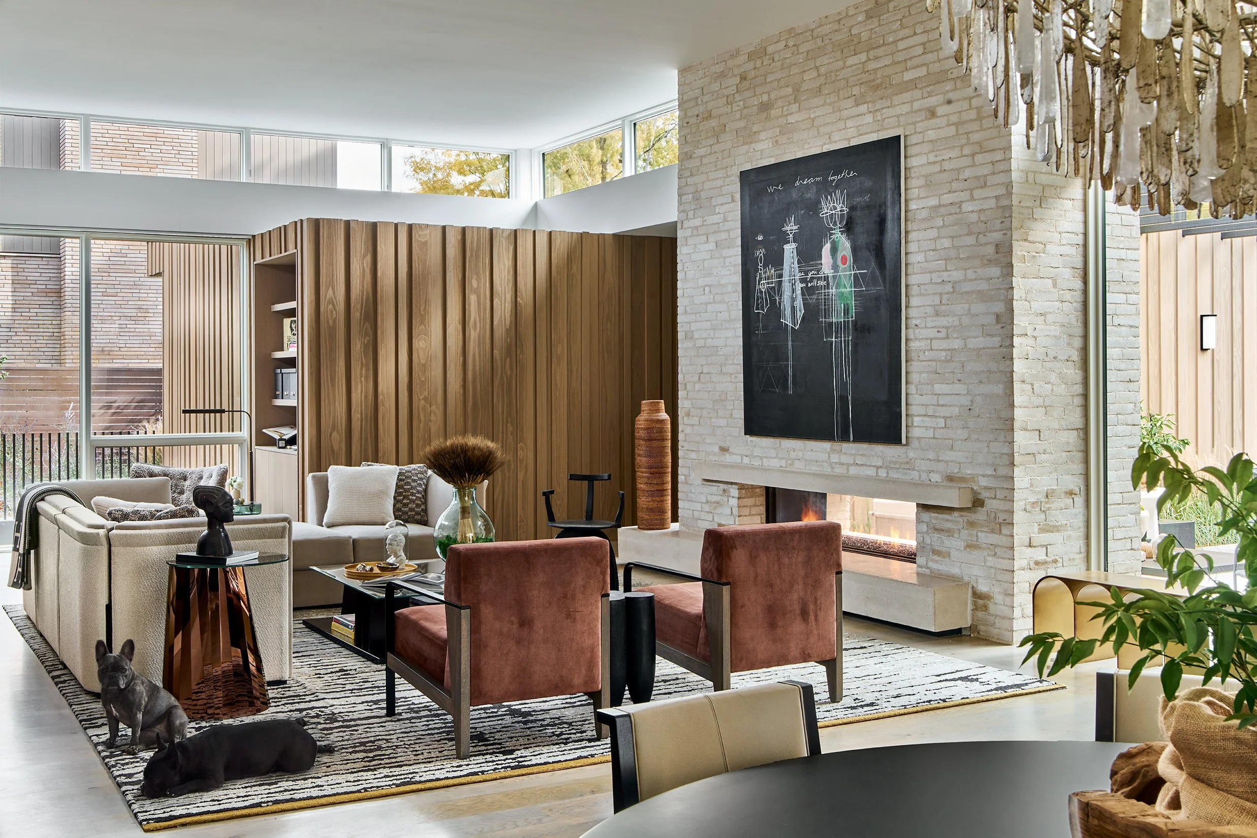 Modern living room with large windows, brick and wood walls, and a white fireplace with a black chalkboard above it. The room has a mix of beige and rust-colored chairs, a rug, and decorative items including a black bust and vases. Two French Bulldogs are seen on the rug, and a chandelier hangs from the ceiling.