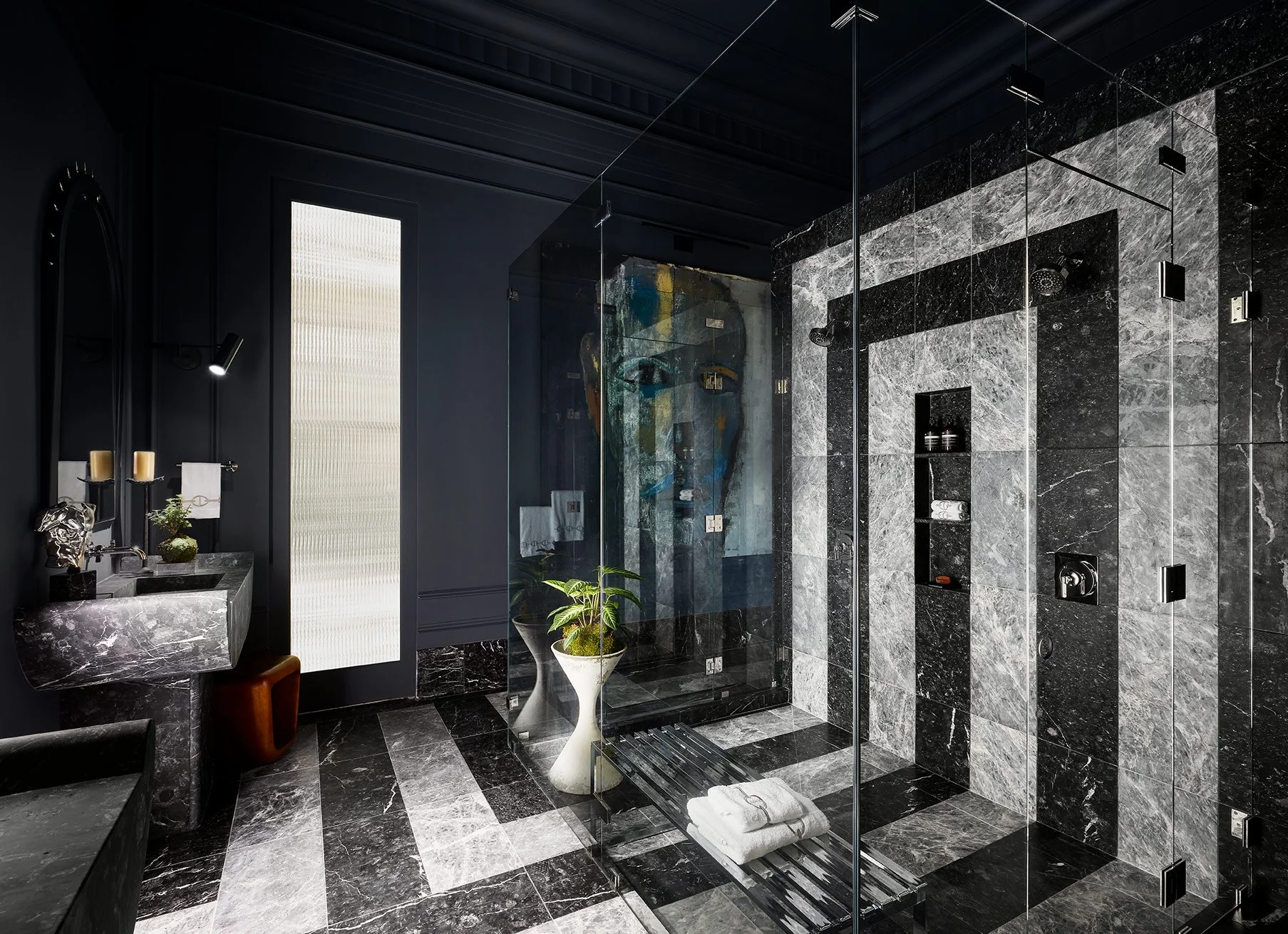 A modern bathroom with black and gray marble tiles, a glass shower enclosure, a floating vanity with a marble countertop, and black fixtures. There is a small potted plant and decorative accessories near the sink, and a textured light panel on the wall.