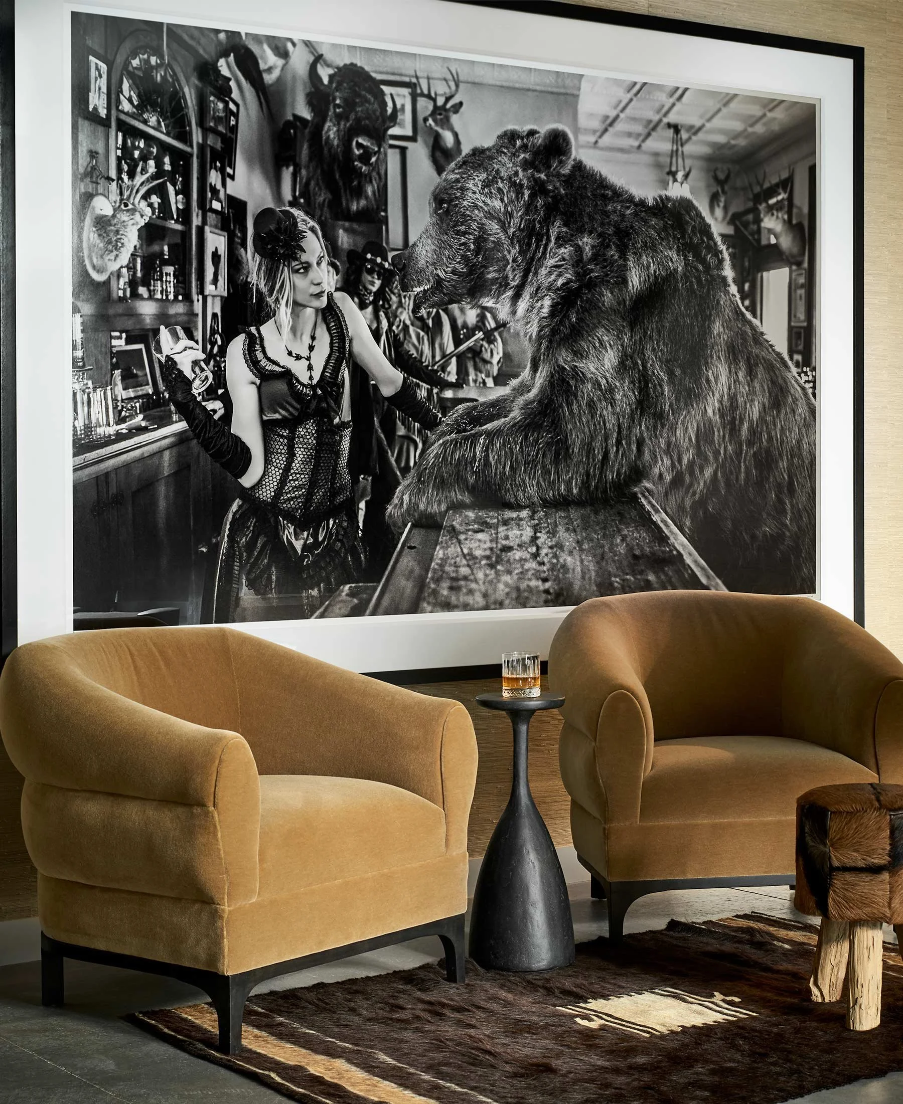 A black-and-white photograph of a bar or lounge with two mustard-colored armchairs, a small black side table with a glass of whiskey, and a large framed picture on the wall depicting a woman dressed in vintage clothing standing next to a bear in a room decorated with mounted animal heads and taxidermy.