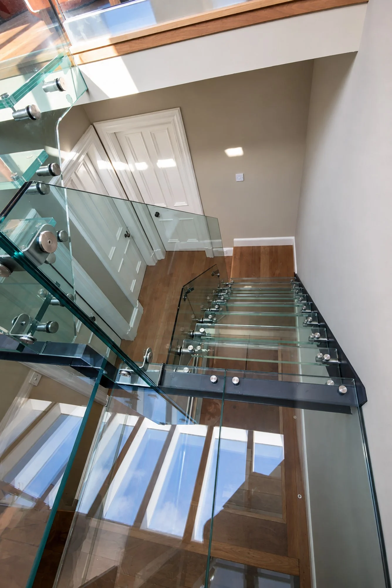 View down through a glazed landing to the glass staircase below, with rooflight reflections across the floor.