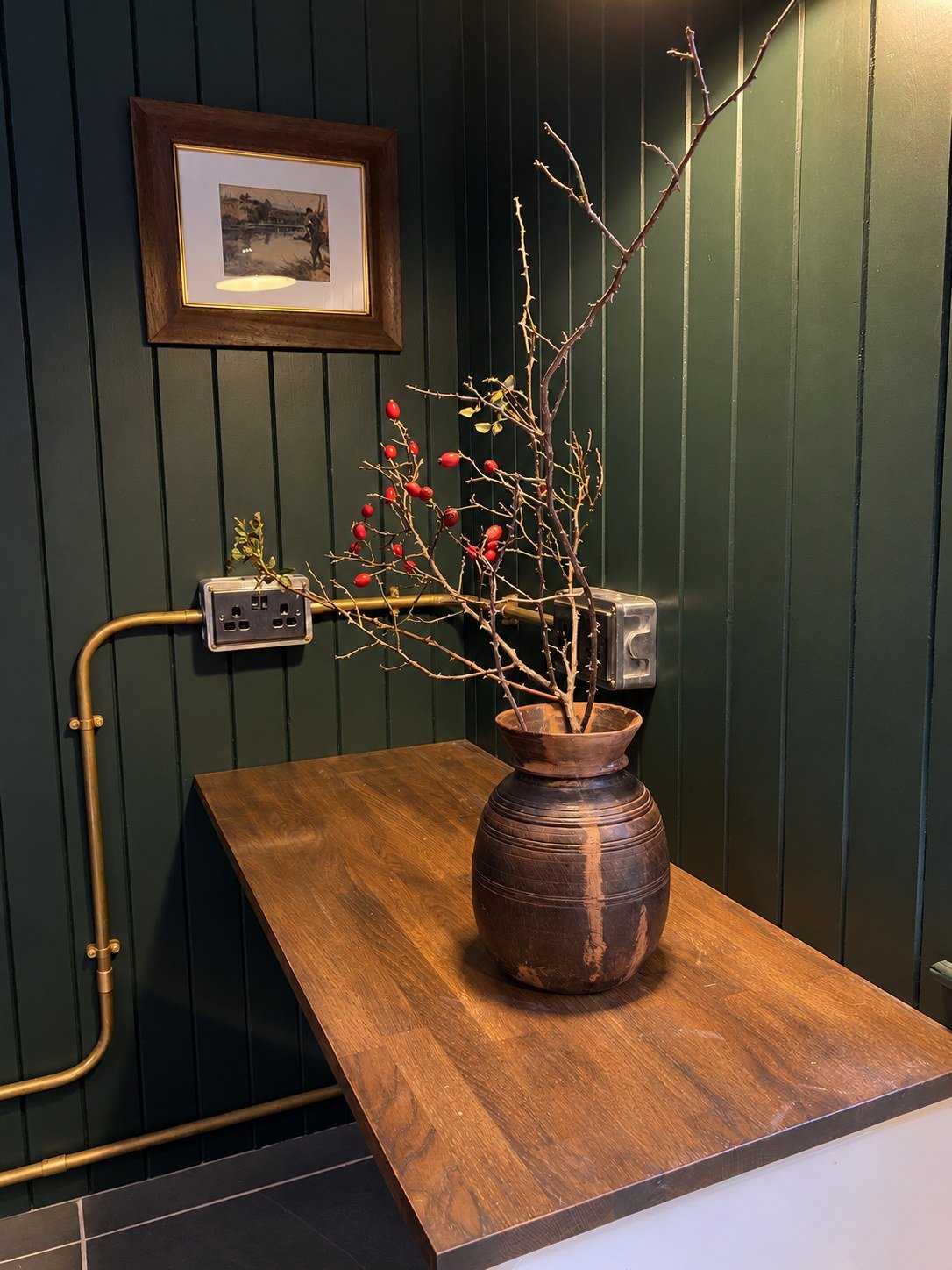 Rosehip branches in a rustic vase beside exposed brass conduit and dark timber panelling in an Aberdeenshire rod room.