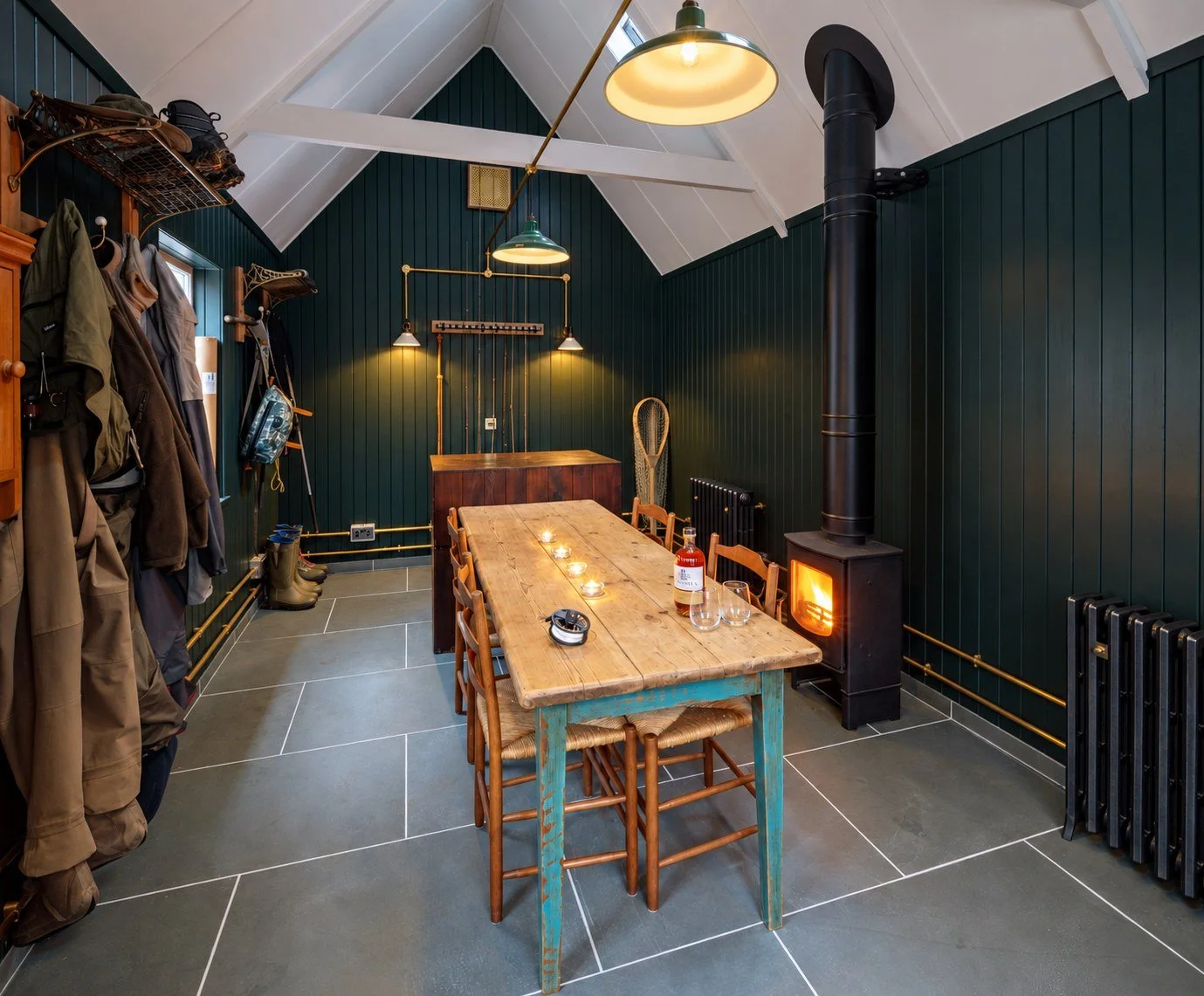 Rod room on an Aberdeenshire sporting estate, with fishing equipment, hanging outdoor clothing, a timber table and a lit wood-burning stove.