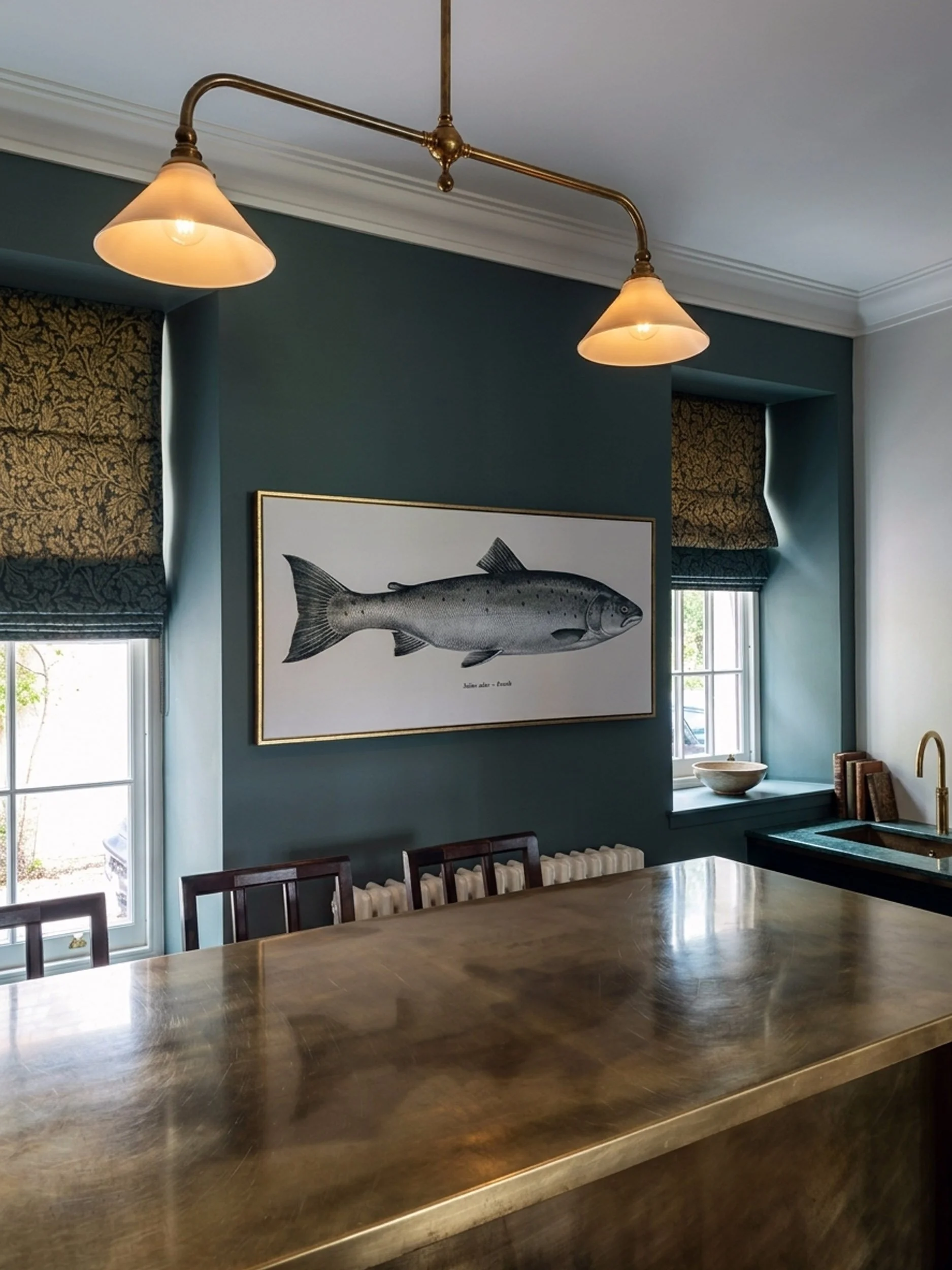 A dining room with a large wooden table, chairs, a blue wall, a framed fish print, and a window with patterned curtains, hanging light fixtures, and a green countertop with a bowl and books.