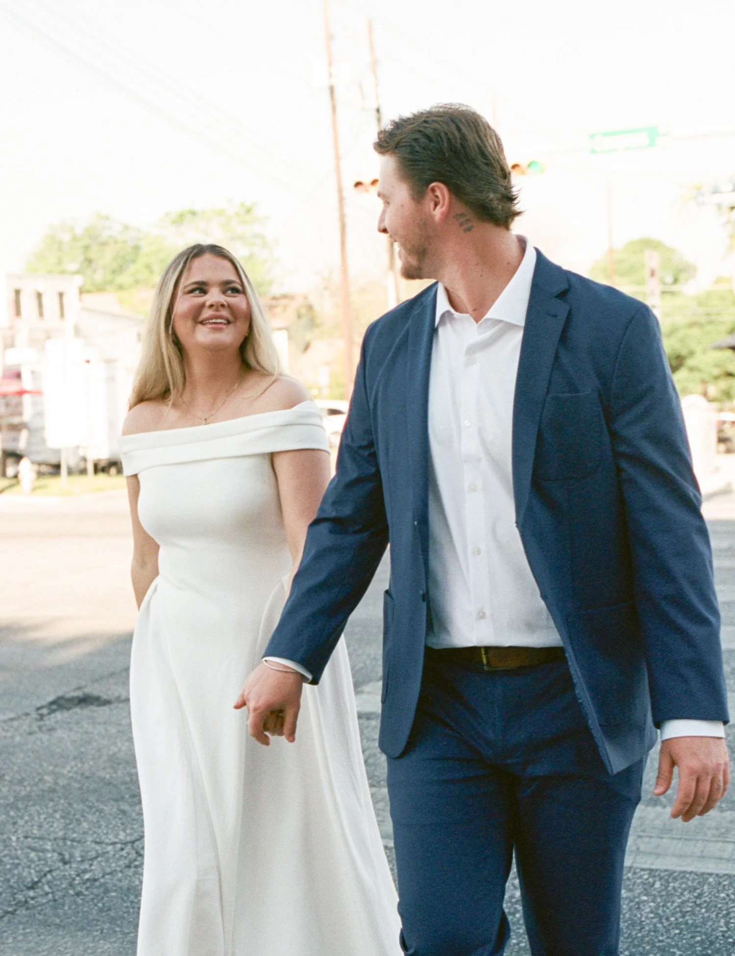  An action shot of wedding couple Brooklyn and Carson as they walk. Carson is slightly in front and is looking back at Brooklyn, while she looks at him. They are both smiling at each other and holding hands. 