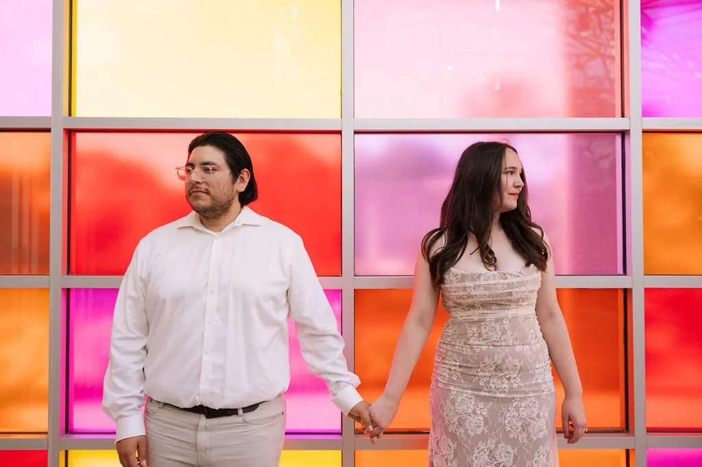 Couple holding hands in front of colorful wall during San Antonio Botanical Garden engagement photos.