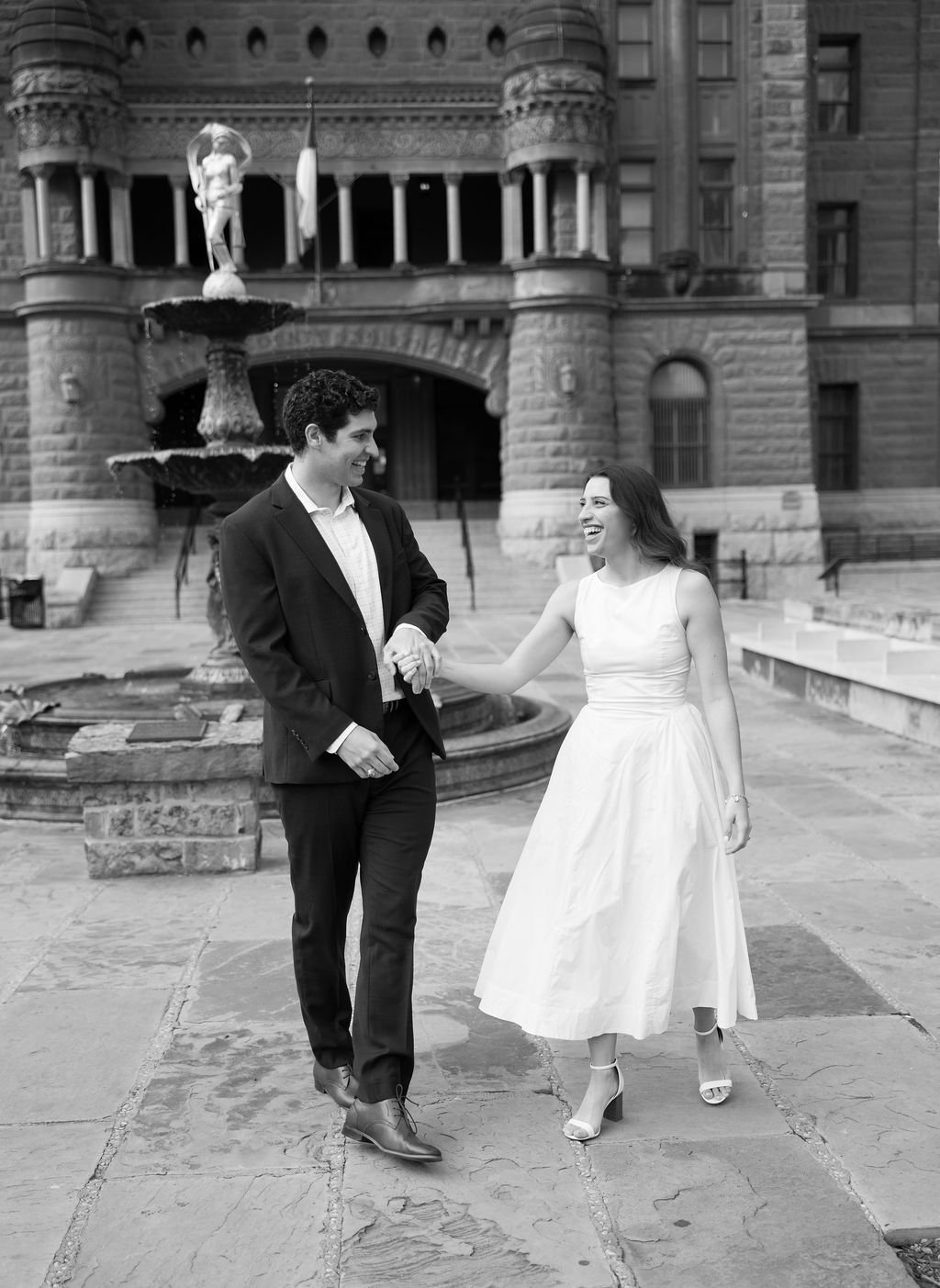 Couple walking hand in hand past fountain during downtown San Antonio engagement photos.
