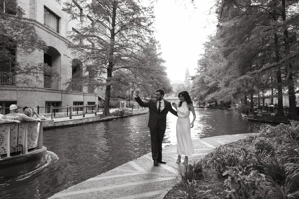 Couple walking along River Walk in black and white, waving to a passing boat tour during San Antonio engagement session.