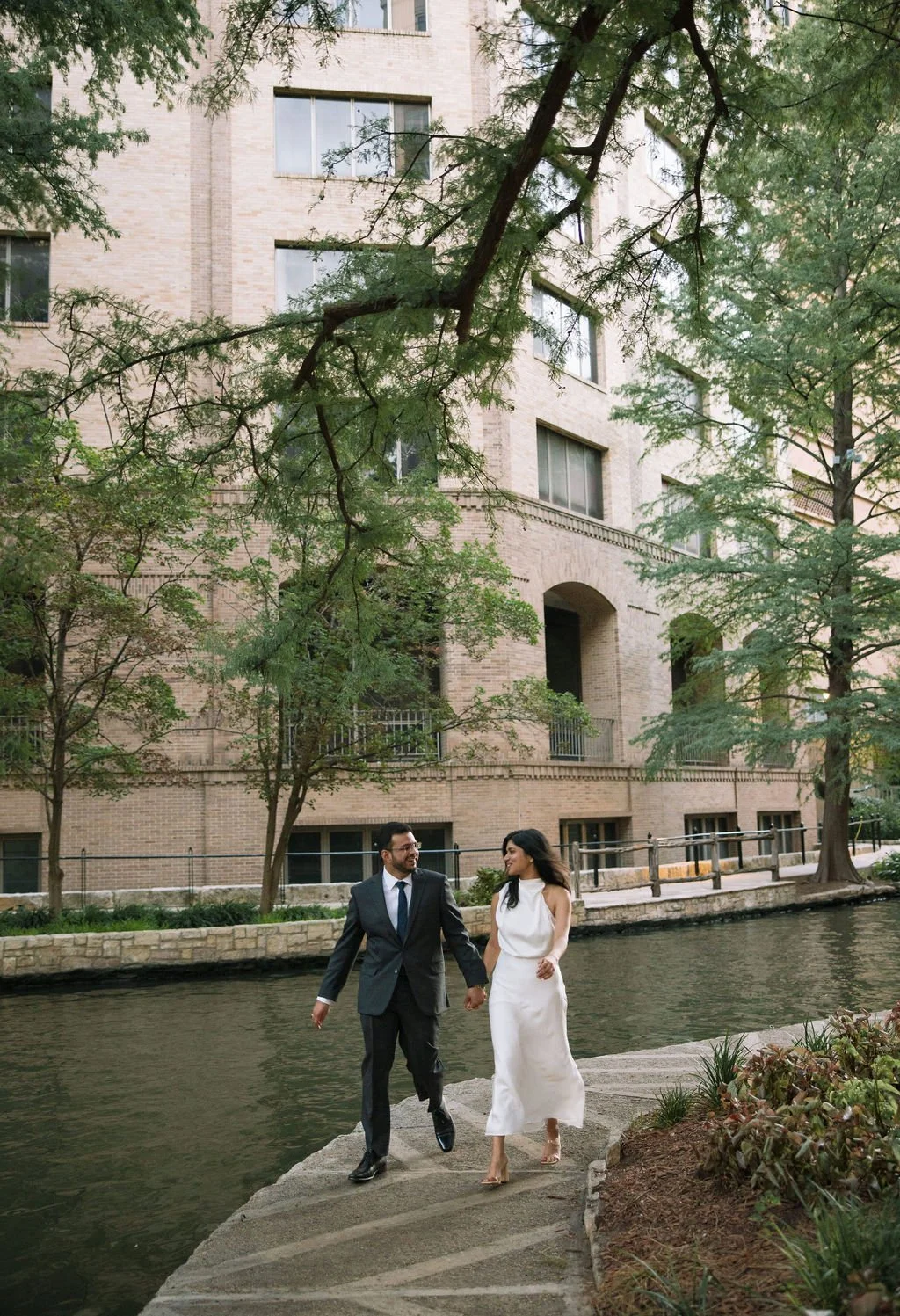 Couple walking together along river path surrounded by trees during engagement photos San Antonio.