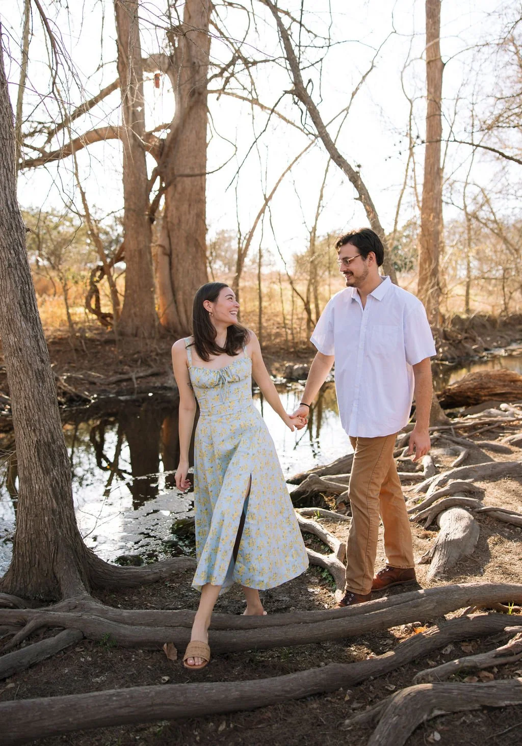 Couple holding hands walking across tree roots at Boerne City Lake Park engagement session.