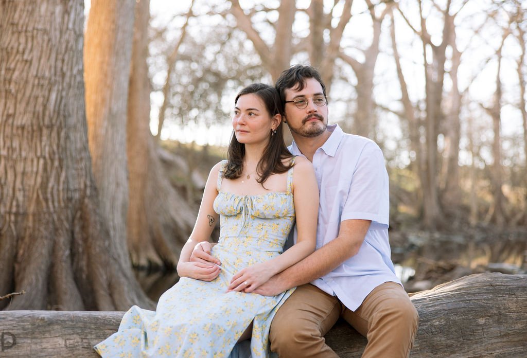 Couple sitting close together on log surrounded by trees during San Antonio engagement session.