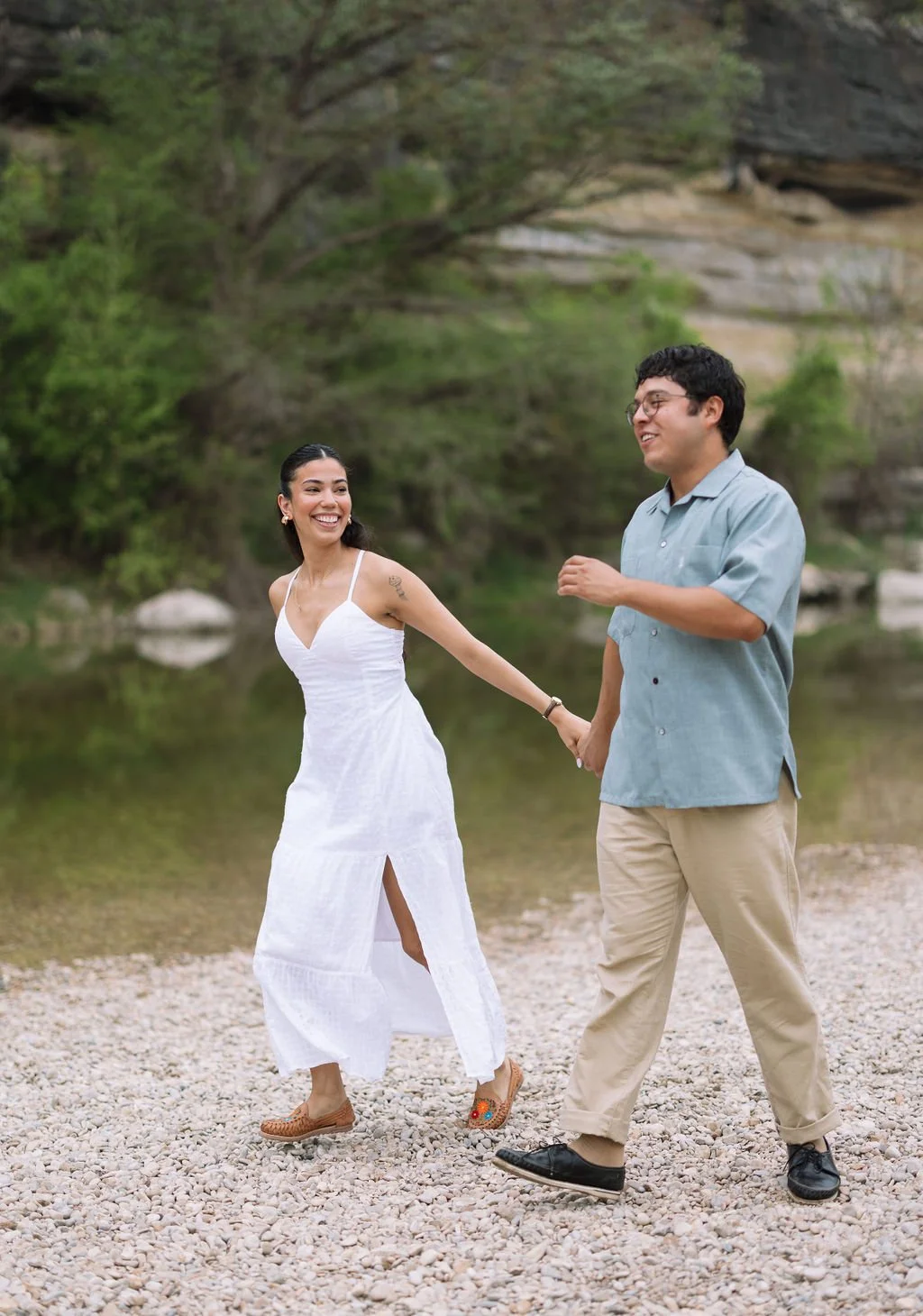 Couple laughing and walking together on riverbank during Guadalupe River State Park engagement photos.
