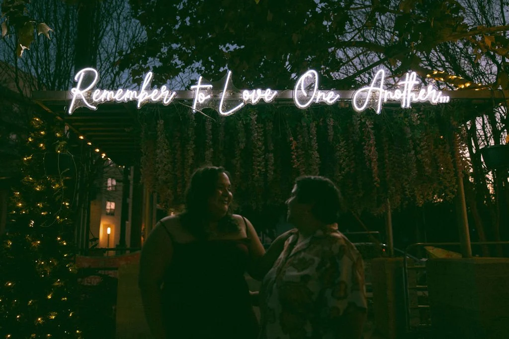 Couple standing under neon sign that reads “Remember to Love One Another” during Idle Beer Hall San Antonio engagement photos.