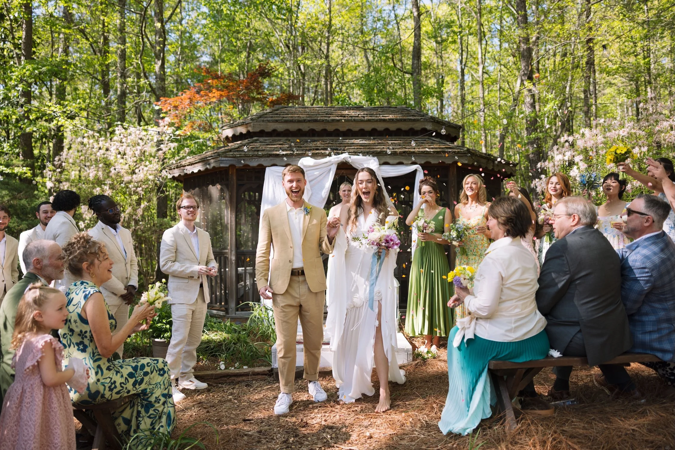A wide shot of bride and groom Lily and Kyle as they just step down the aisle, post-ceremony. They have wide open smiles on their faces as they are surrounded by their cheering and clapping loved ones.