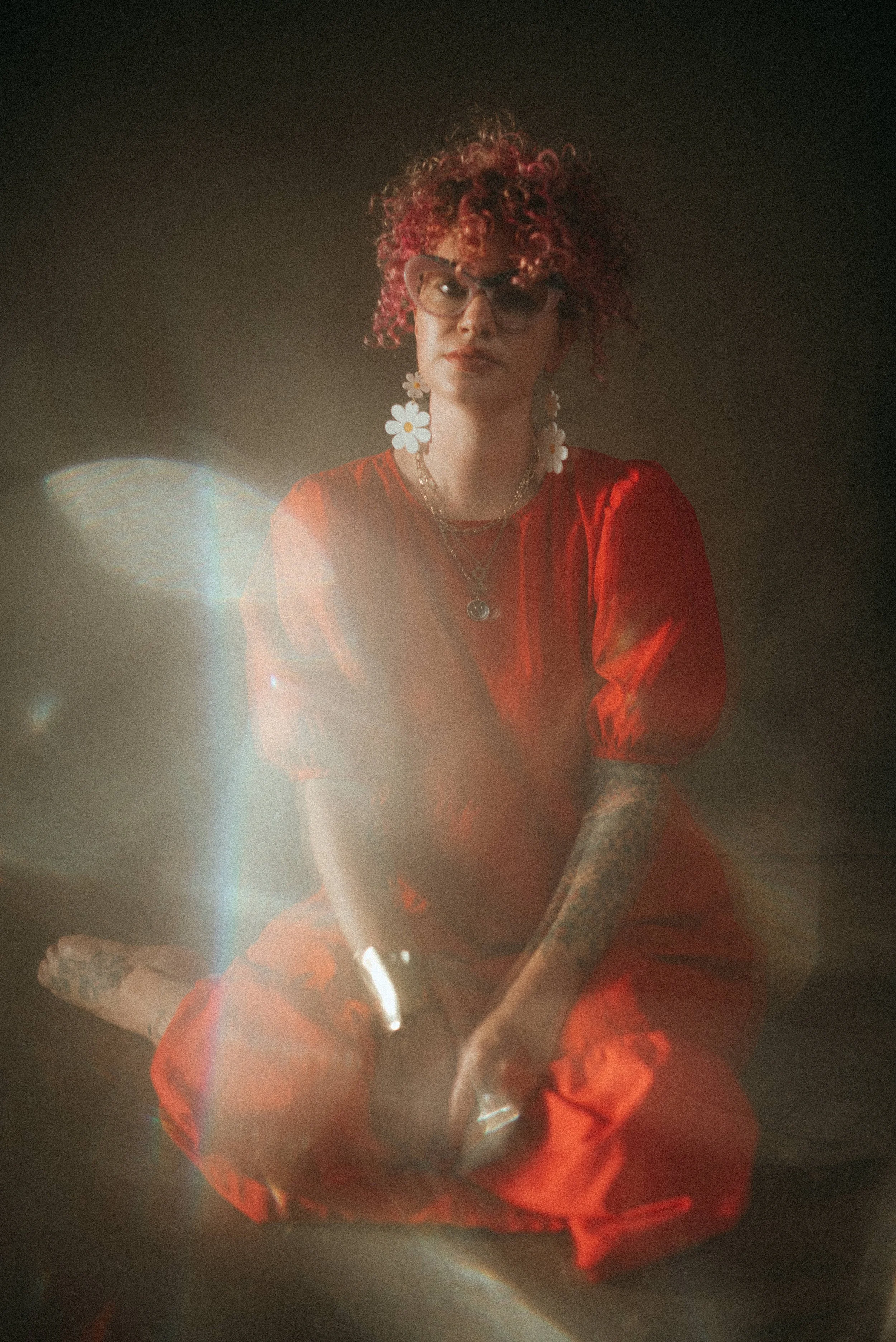 Documentary style photographer, Christy Ann, sits on a floor with a dark backdrop behind her. She has curly red hair, pink cat eye glasses, large daisy earrings, and wears a bright red dress.