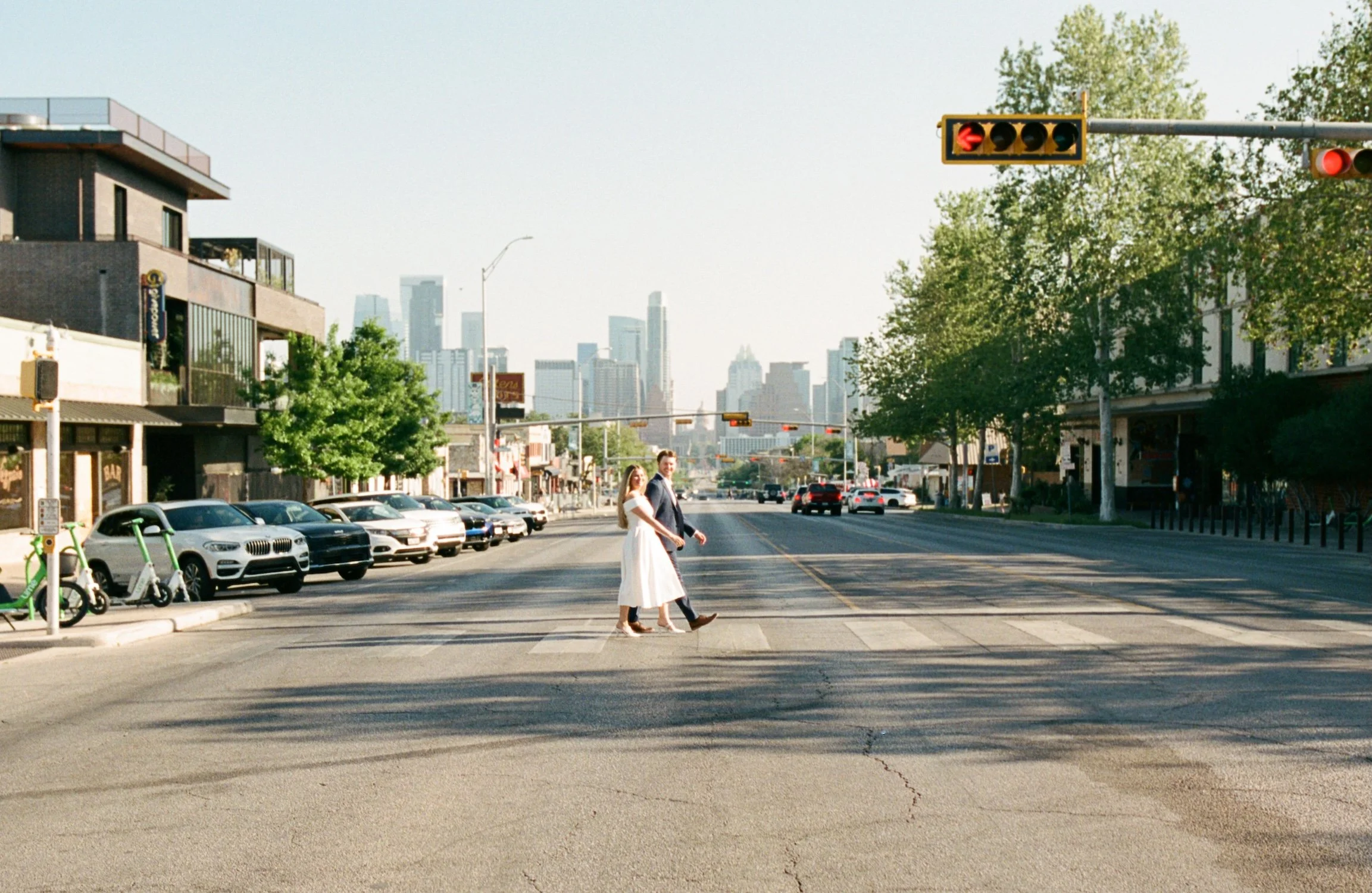  A distance shot of Brooklyn &amp; Carson walking across an Austin, TX street. They are centered in the photo and are walking side by side as they smile at the camera. Smaller business shops can be seen on either side of the street, while the Austin 