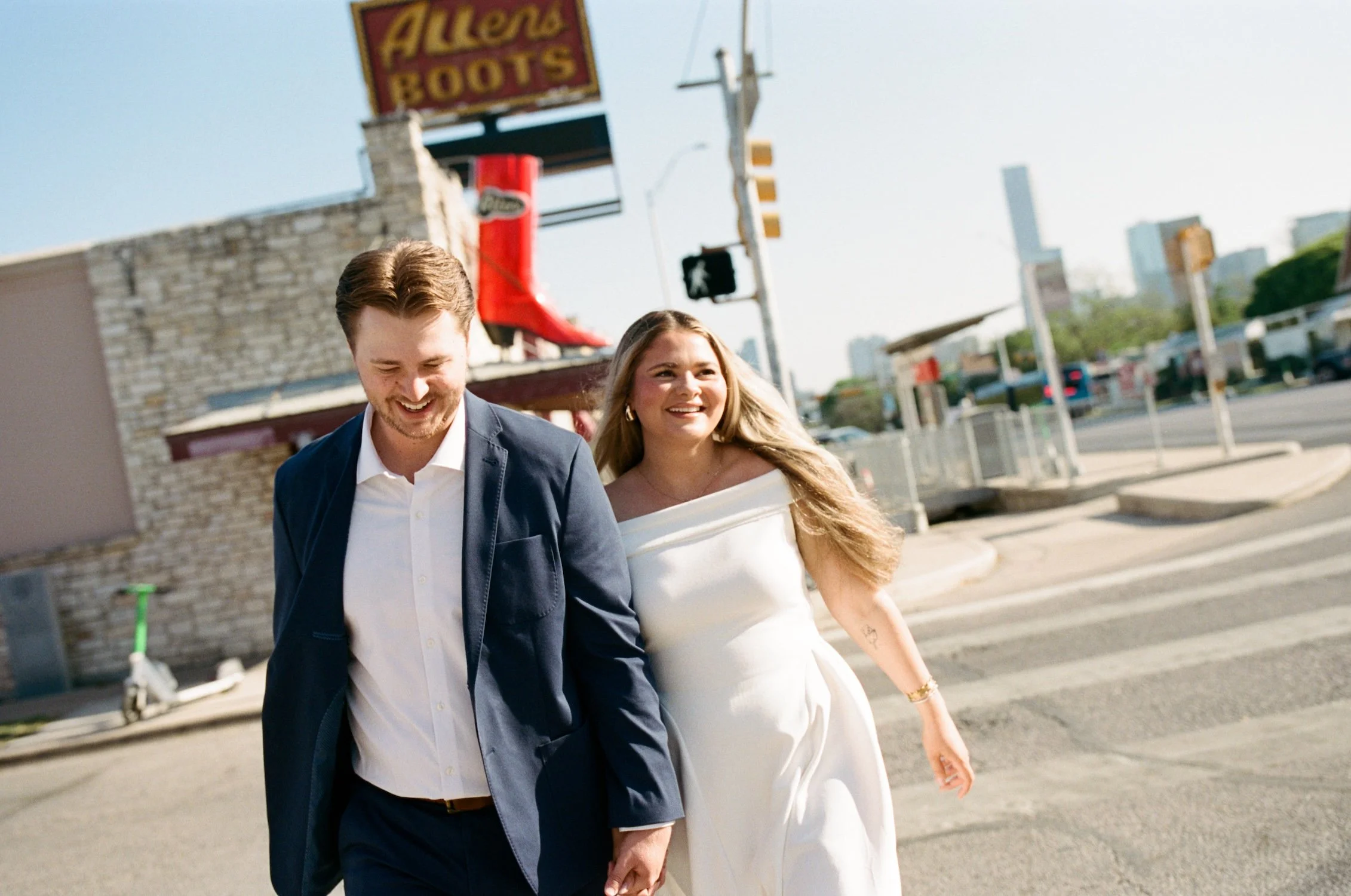  An action shot of Carson and Brooklyn as they walk across the street. Carson is slightly in front and is looking down while smiling. Brooklyn is looking off to the right white smiling. A cowboy boot store sign can be seen just over their heads. 