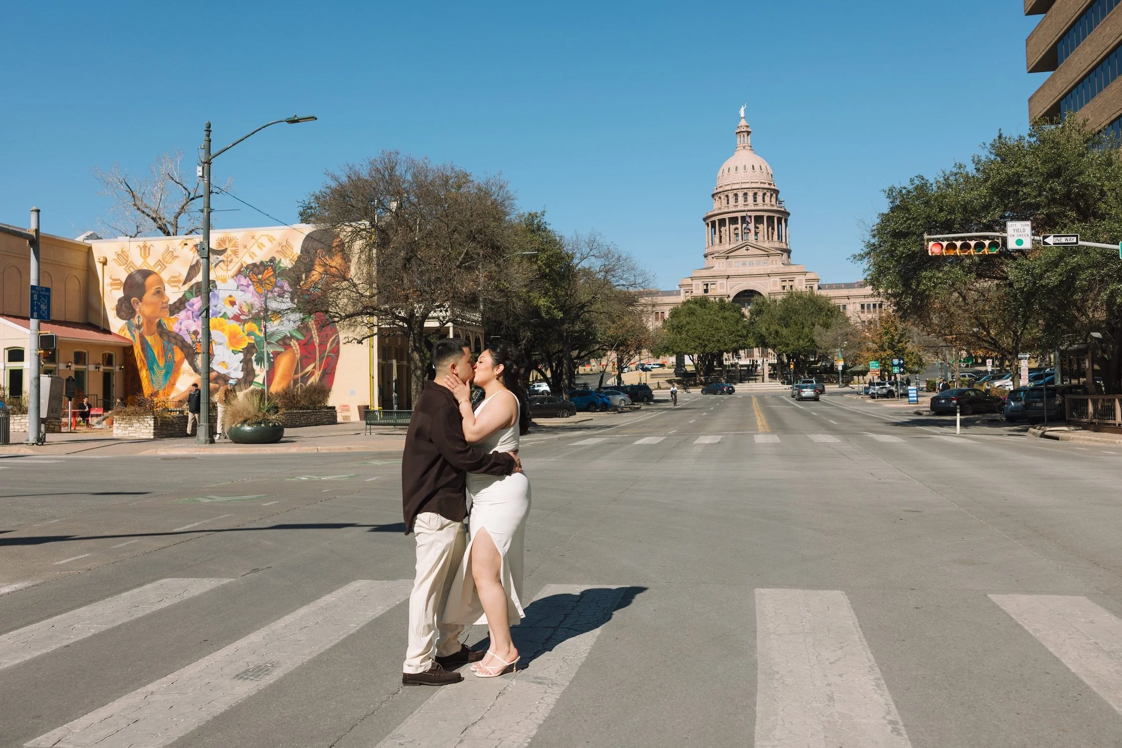 Savannah & Dominic, both of apparent Latin descent, stand in the middle of a street while kissing and embracing. A colorful mural on a building side and the Austin, TX Capitol building can be seen in the background.