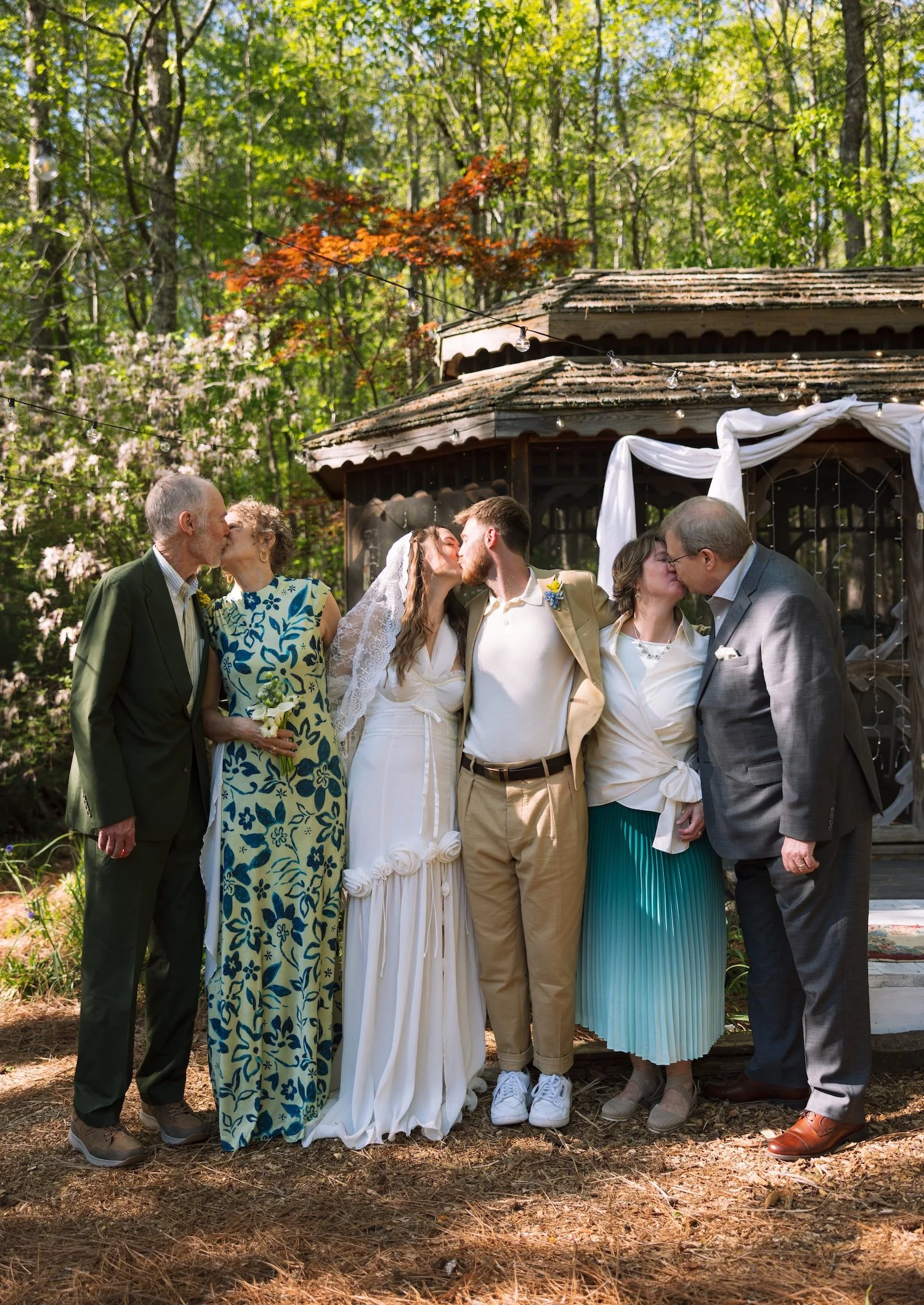 Lily and Kyle are centered while each set of their parents stand on either side of them. All three couples stand arm in arm while turned towards their partner in a kiss.