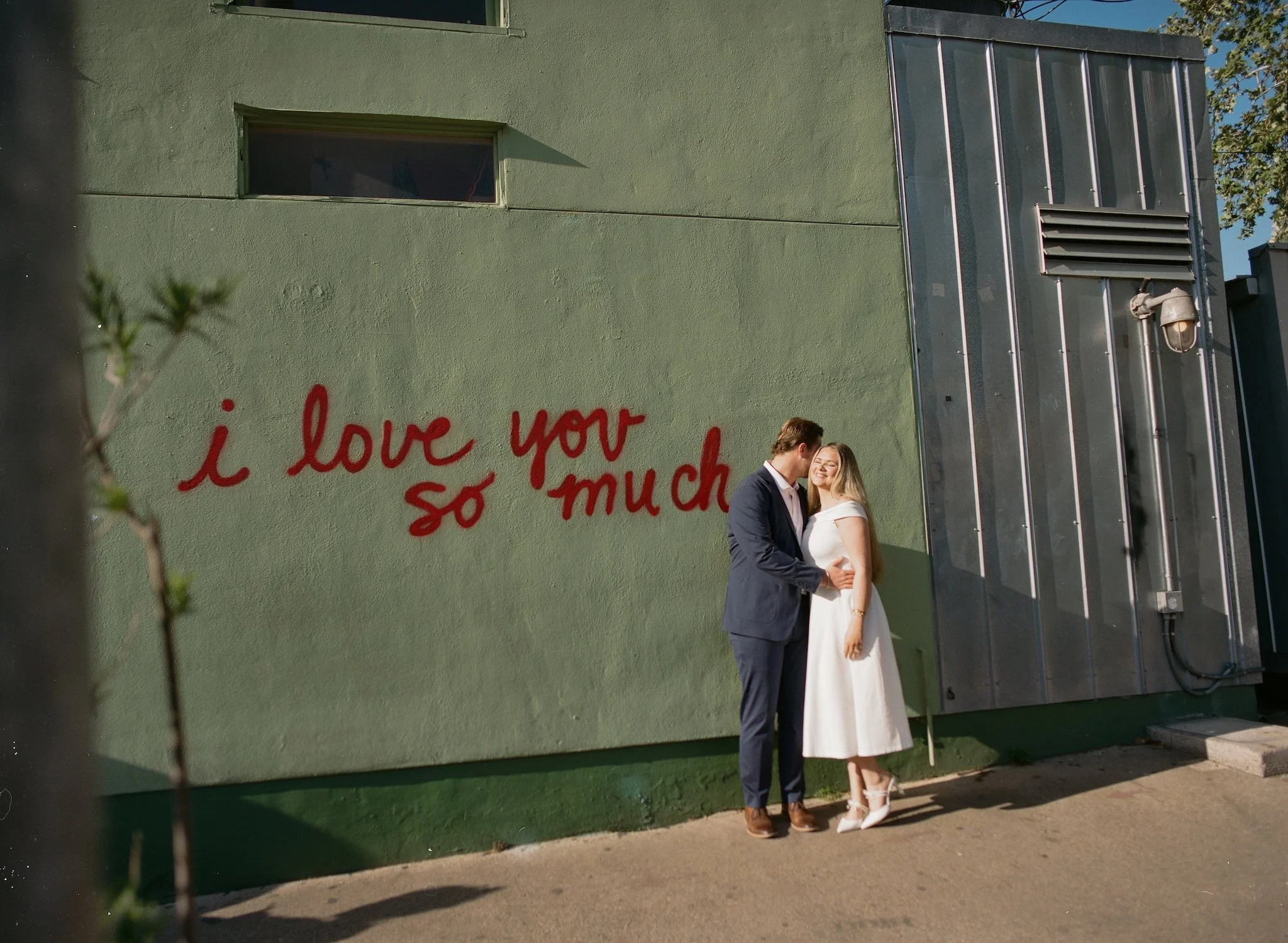 Brooklyn & Carson, both white, are embracing while standing in front of an Austin, TX mural. The mural is of simple red cursive text that says, "i love you so much," on a sage green wall.