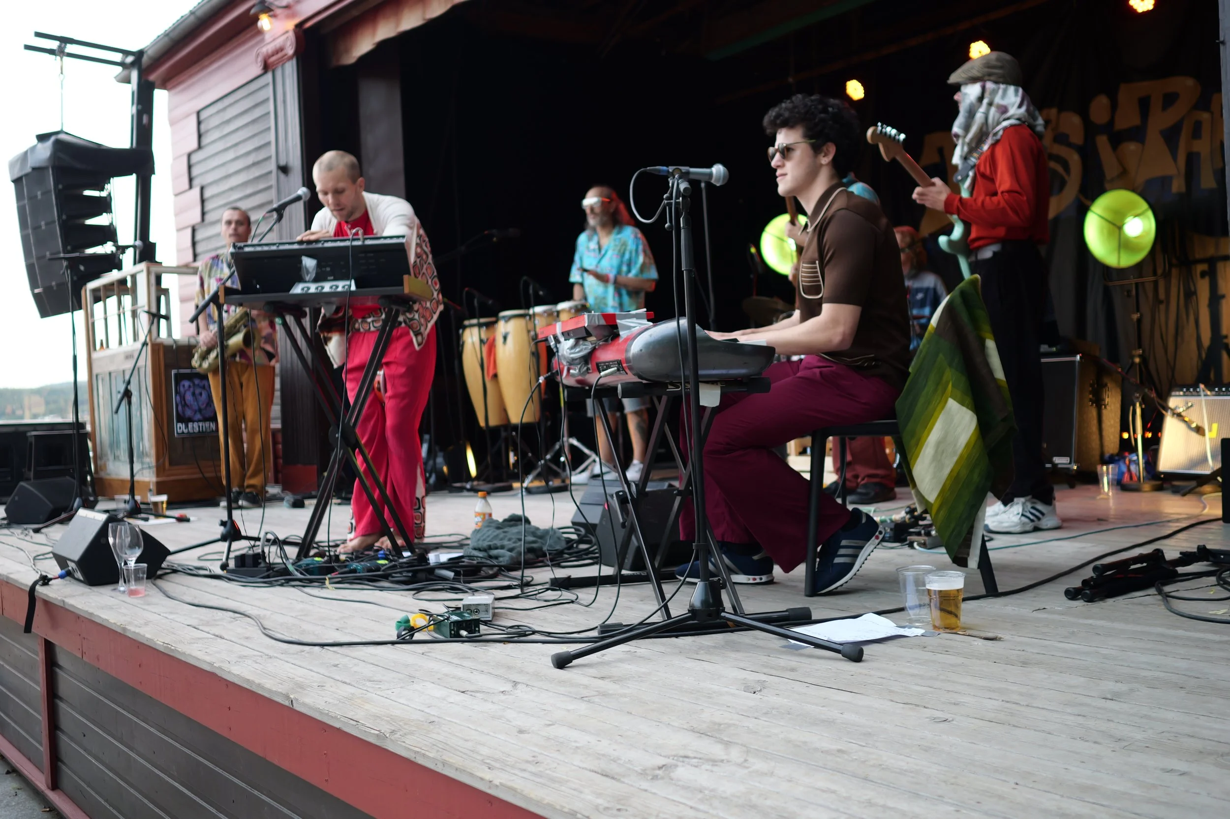 Et band på en utendørs scene spiller musikk, med flere instrumenter som keyboard, perkusjon og gitar. Musikerne er kledd i fargerike klær, og scenen er utstyrt med lyd- og lysutstyr.