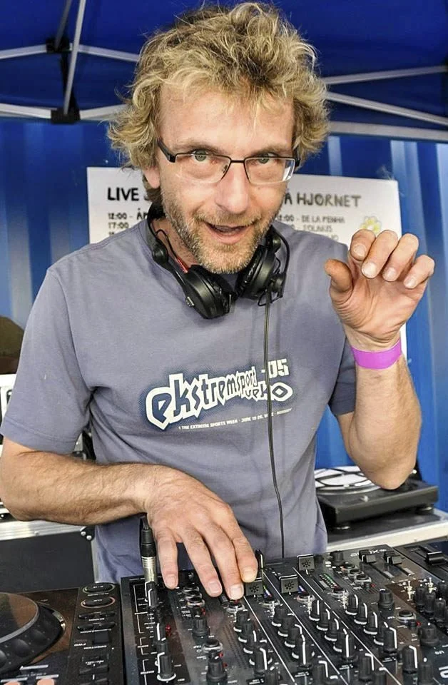 En mann med briller og lyst, krøllete hår som står bak en DJ-konsoll, gjerne kjent som DJ, med hodetelefoner rundt halsen og et blått telt i bakgrunnen.