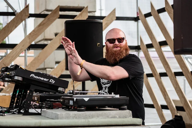 En mann med rismaske og bart, iført solbriller, står bak DJ-utstyr på en scene, ser mot kameraet og smiler.
