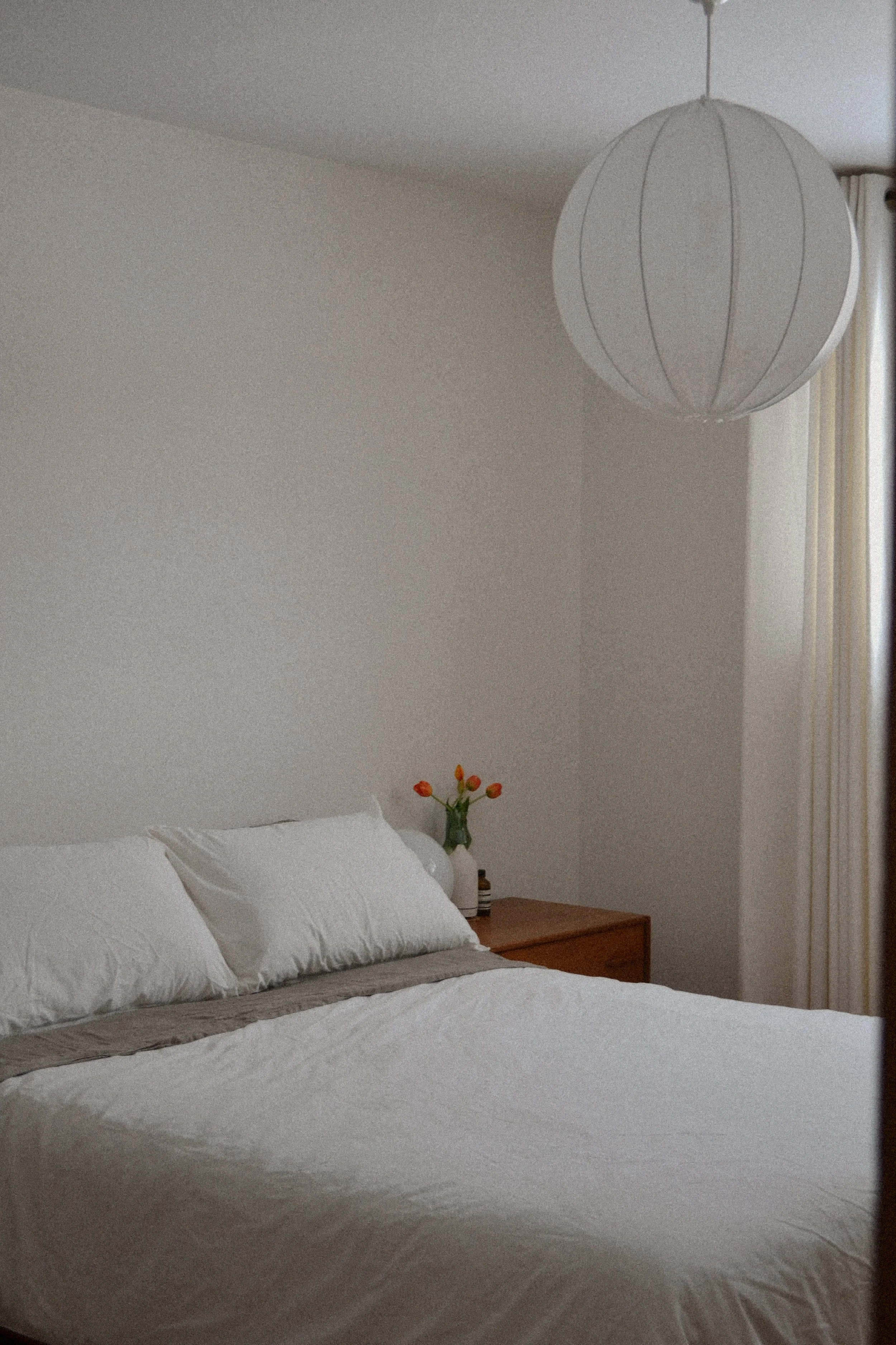 Minimalist bedroom with white bedding, a wooden nightstand, vase of orange tulips, and a round paper lamp hanging from the ceiling.