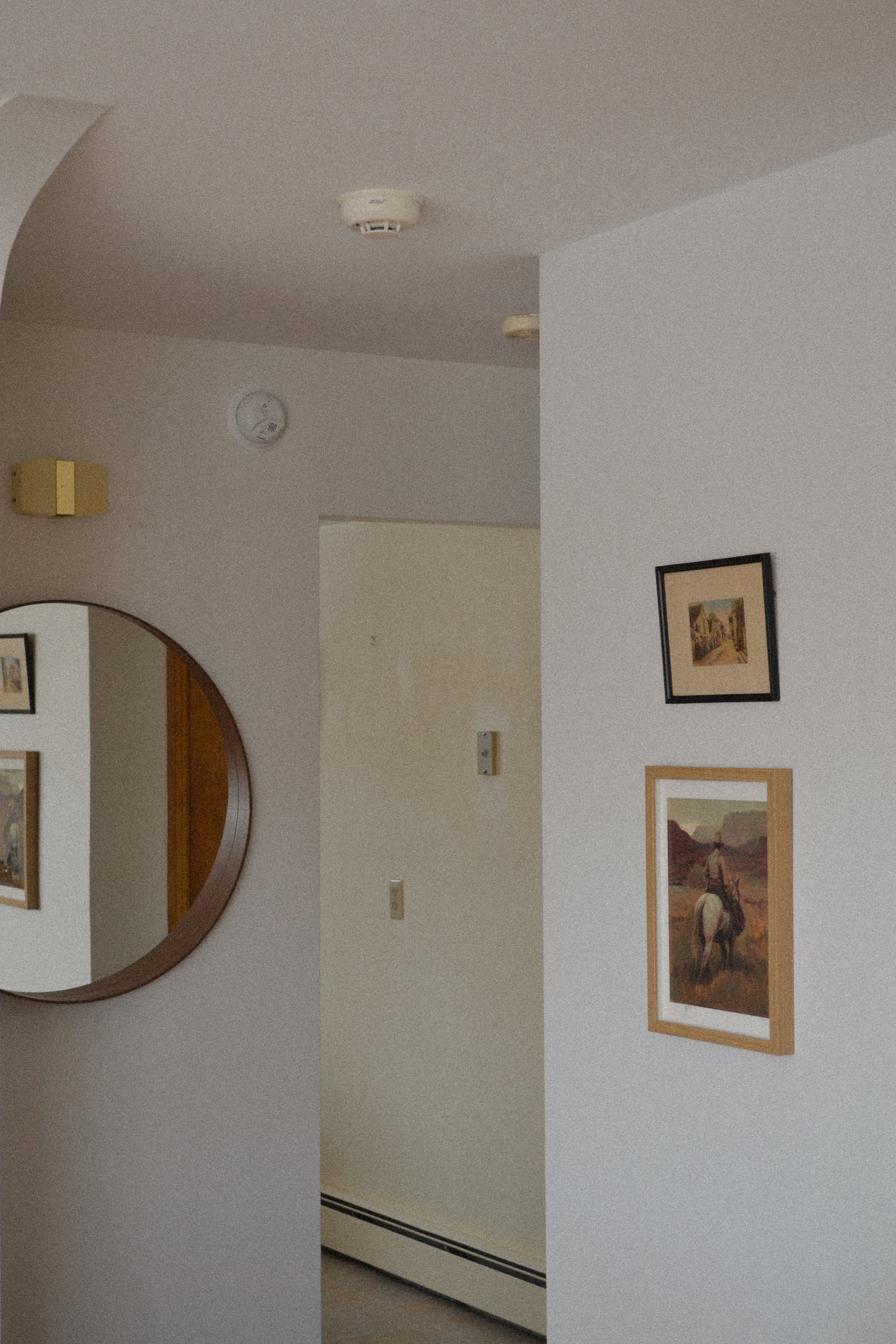 Interior of a room with a white wall, round mirror, framed artworks, smoke detector on the ceiling, and a light fixture.