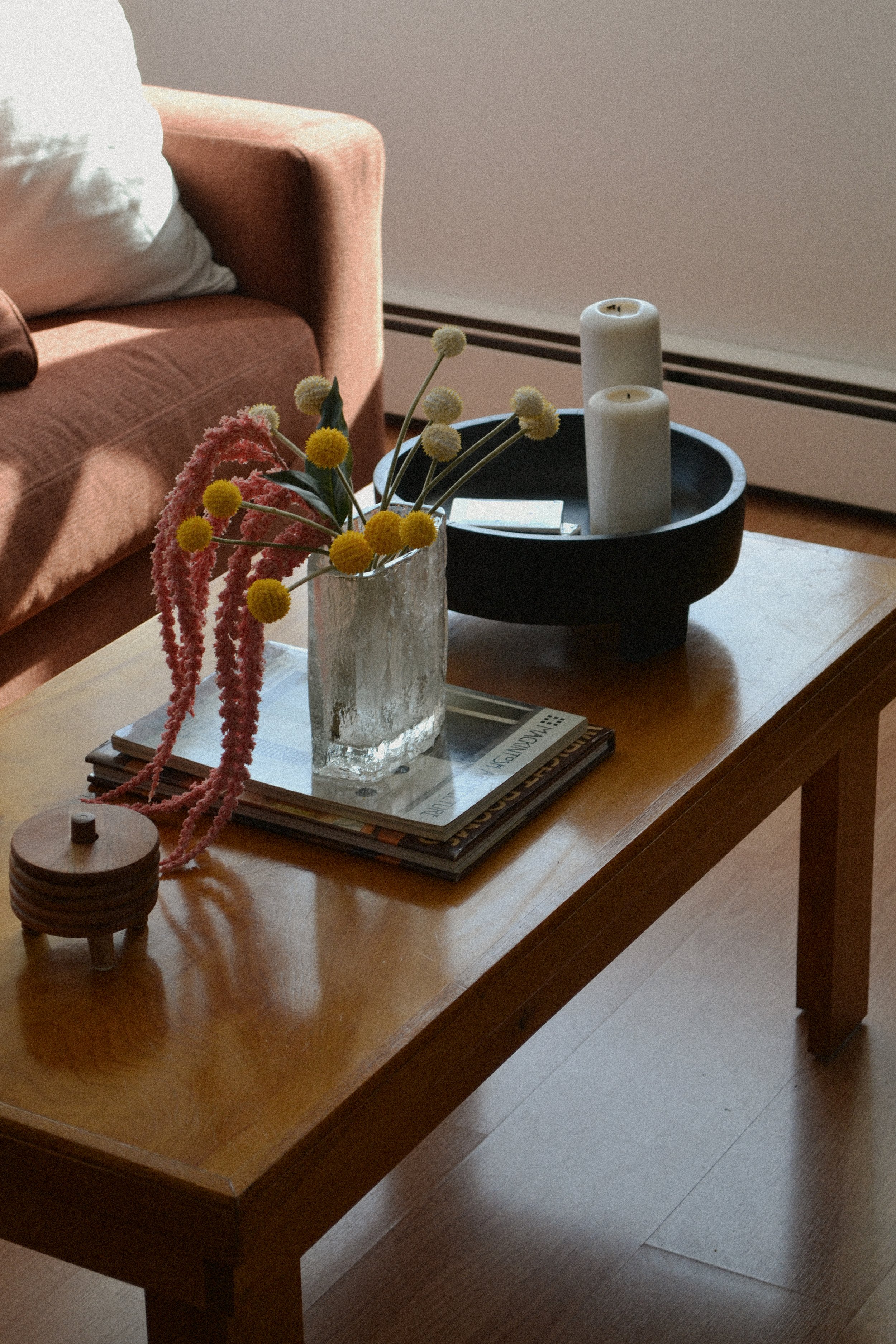 Living room scene with brown couch and rustic wooden coffee table. Table features glass vase with pink and yellow flowers, stacked magazines, round wooden container, and tray with two white candles.
